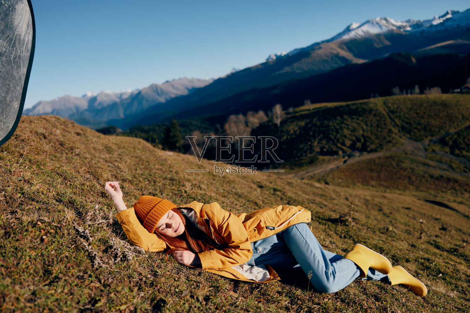 女人躺在山上的草地上休息,微笑着用牙齿看着山上的雪在冬天穿着黄色的雨衣和牛仔裤快乐的日落旅行徒步旅行,自由的生活方式照片摄影图片