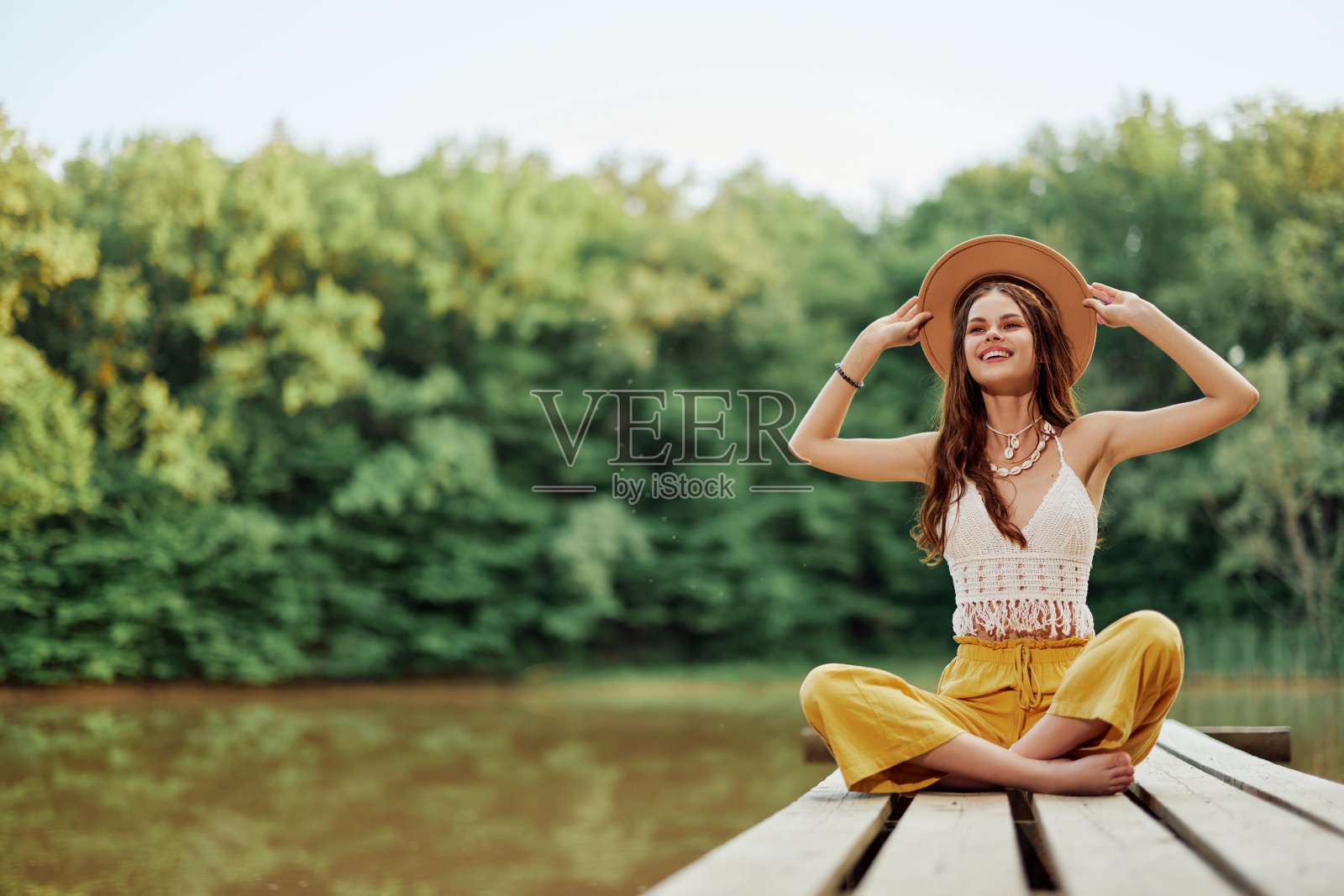 一位嬉皮生态活动家坐在湖边的桥上,她伸出双臂,戴着帽子,真诚地微笑着照片摄影图片