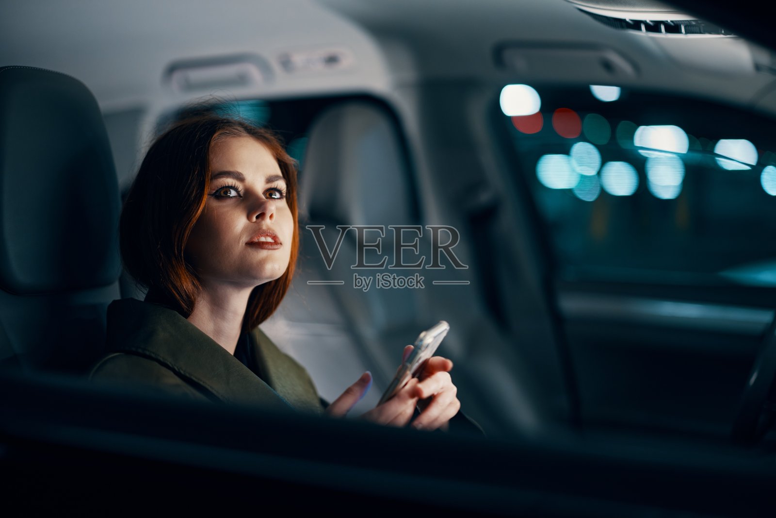 这是一幅近距离的横向肖像,一位穿着皮衣的时尚、奢华的女子晚上坐在一辆黑色轿车的副驾驶座位上,若有所思地向开着的车窗外望去照片摄影图片