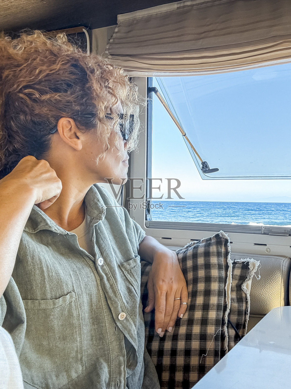 戴眼镜的卷发女人微笑地喝着茶,望着她的露营车窗外。她在车轮上度过居家假日,享受海边的春季冒险。游牧生活方式概念照片摄影图片