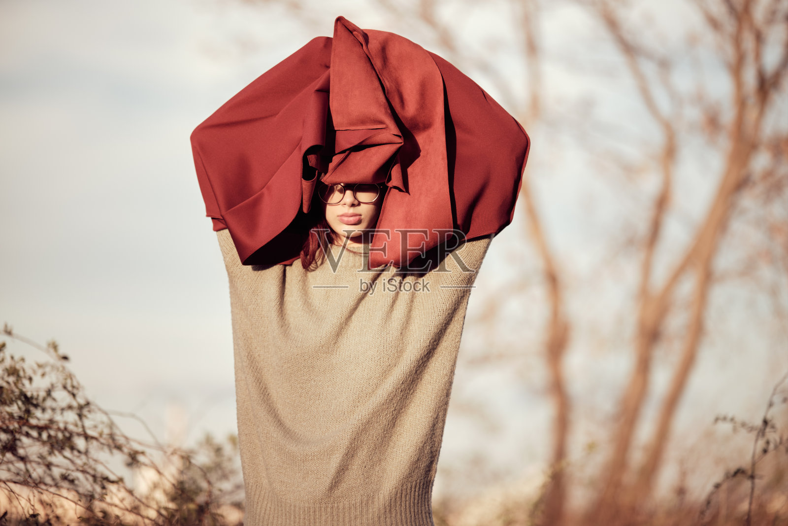 冷静,严肃的十几岁的女孩站在公园里,双臂举过头顶,红色的外套遮住了脸的上部和眼睛。照片摄影图片