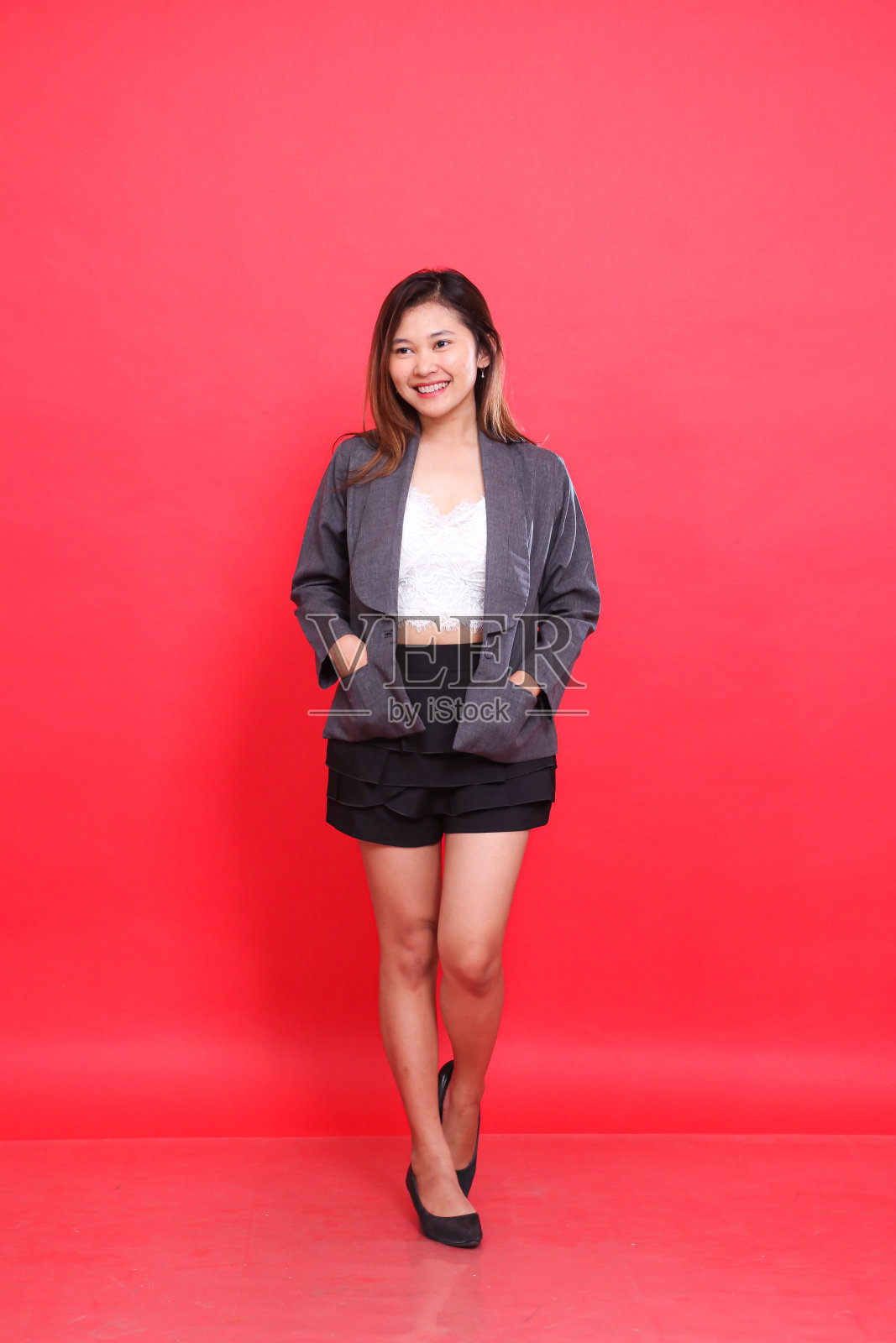 身材丰满的亚洲女孩穿着灰色西装,坦率地微笑着走着,长发,白色的皮肤在红色背景的工作室里。用于企业概念、时尚和广告照片摄影图片