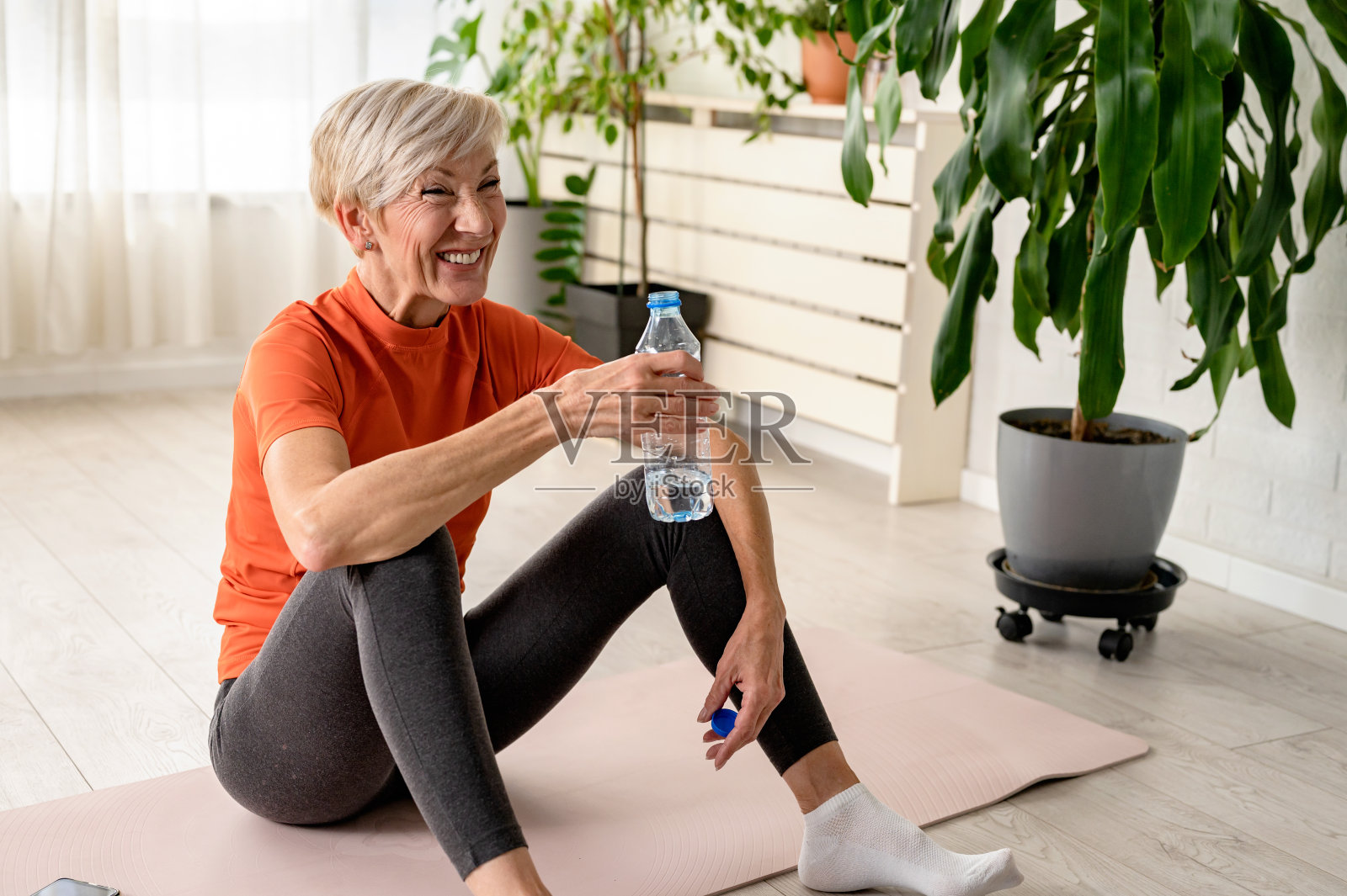 漂亮的老女人运动后喝水。活跃的资深女性在室内练习瑜伽。老年人的运动照片摄影图片