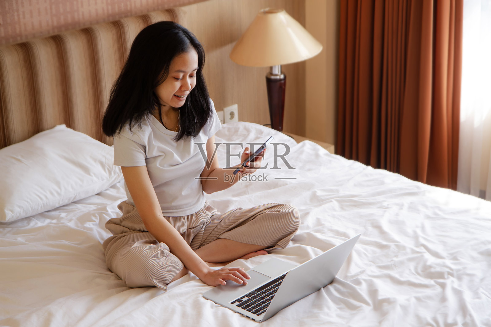 快乐的亚洲女人在家里的床上用笔记本电脑和智能手机照片摄影图片