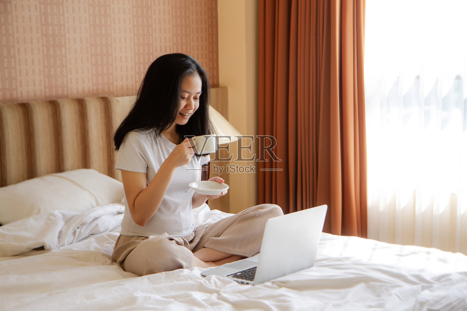 女人在床上悠闲地喝着咖啡,玩着笔记本电脑。快乐的年轻亚洲印尼女人在家里躺在床上放松笔记本电脑。照片摄影图片