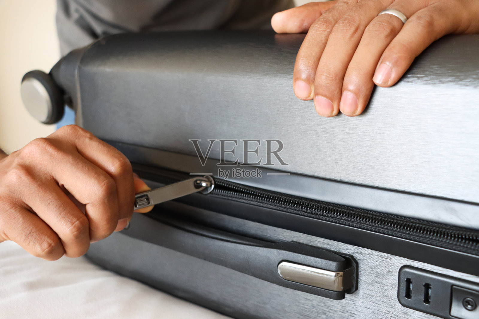 灰色的轮式行李箱放在双人床上,上面盖着白色羽绒被,一个不认识的男人俯身拉上行李的拉链,密码锁,旋转的圆盘,三位数的数字序列,聚焦在前景照片摄影图片