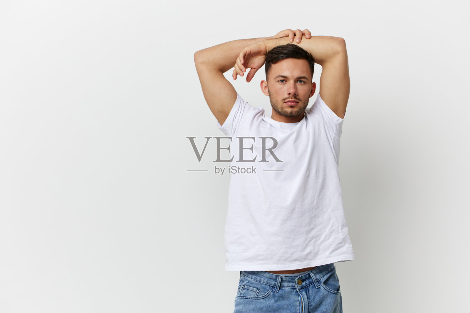 年轻、轻松、快乐、皮肤黝黑的英俊男子穿着基本款t恤,双手交叉在头顶,在白色的摄影棚背景上单独摆姿势。复制空间横幅模型。人的情感生活理念照片摄影图片