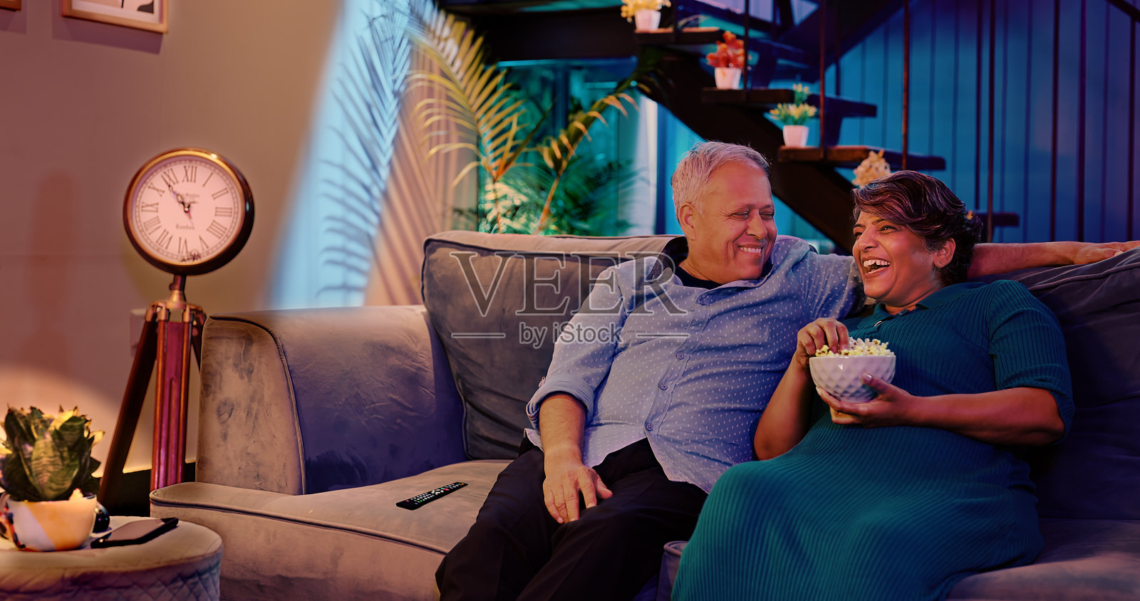 幸福的印度老年夫妇坐在舒适的沙发上,在家里一起看电视上的喜剧电影。年老退休的丈夫、妻子、奶奶、爷爷在家里吃着爆米花、拿着碗、看着搞笑电影照片摄影图片
