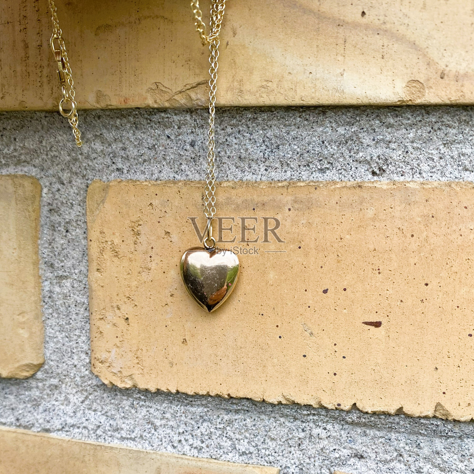 一条链子上挂着一个金色的心形吊坠,靠在黄色的砖墙上。照片摄影图片