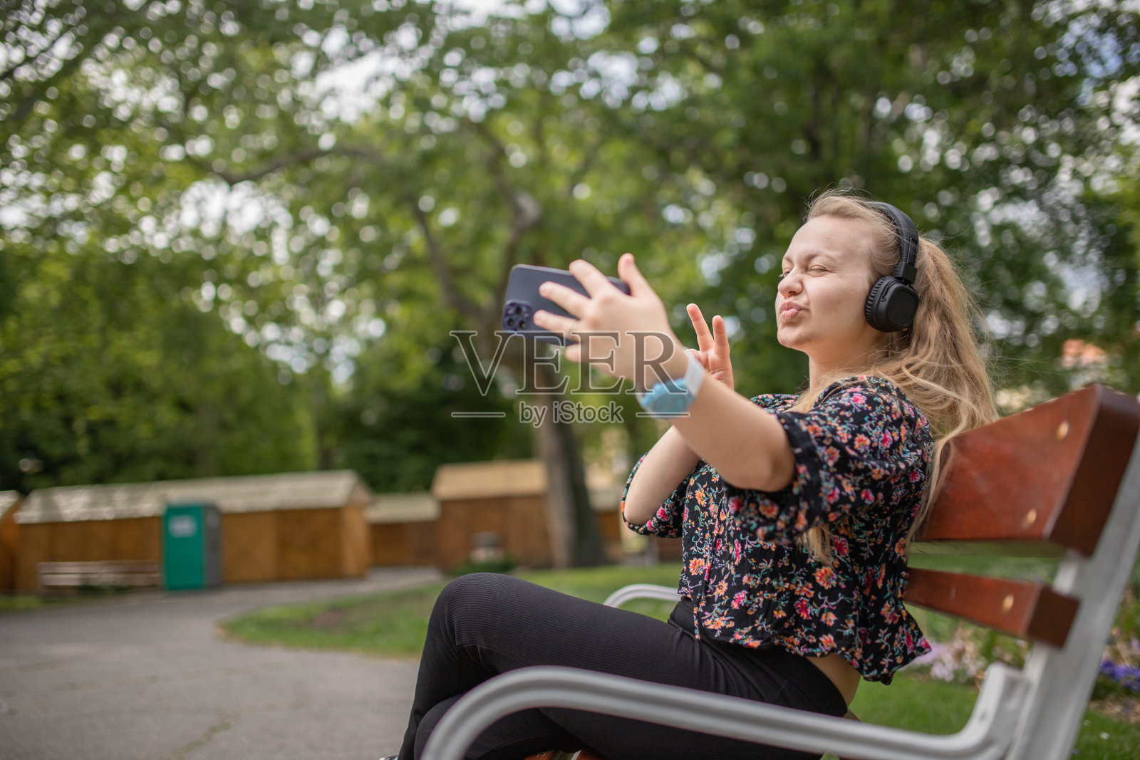 一名年轻女子坐在公园的长椅上,一边用智能手机,一边做鬼脸,一边自拍照片摄影图片