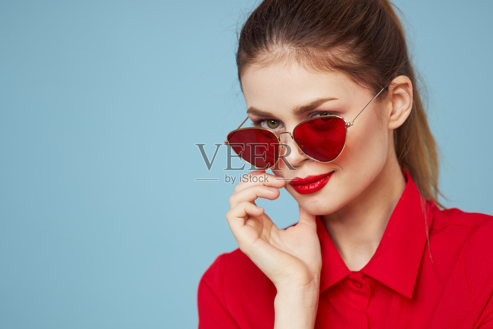 美丽的女人戴着墨镜,红衬衫,明亮的妆容,情绪迷人,蓝色的背景照片摄影图片