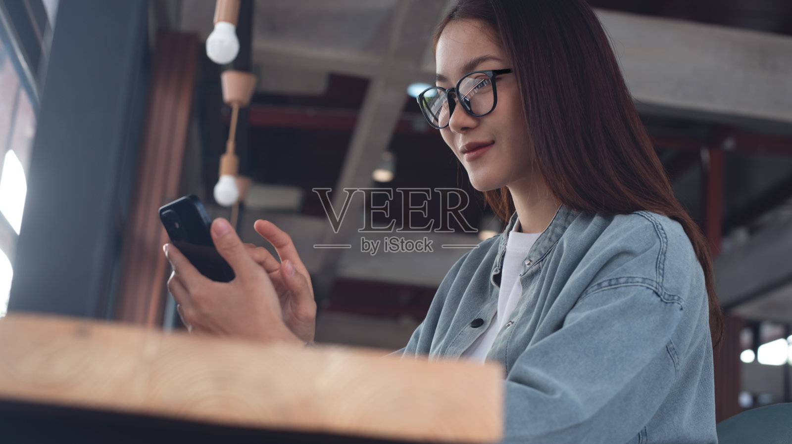 一位戴眼镜的亚洲女性坐在咖啡店的桌子上,用着手机。微笑的年轻女子在咖啡馆看着智能手机和在线消息,社交媒体网络,现代人的生活方式照片摄影图片