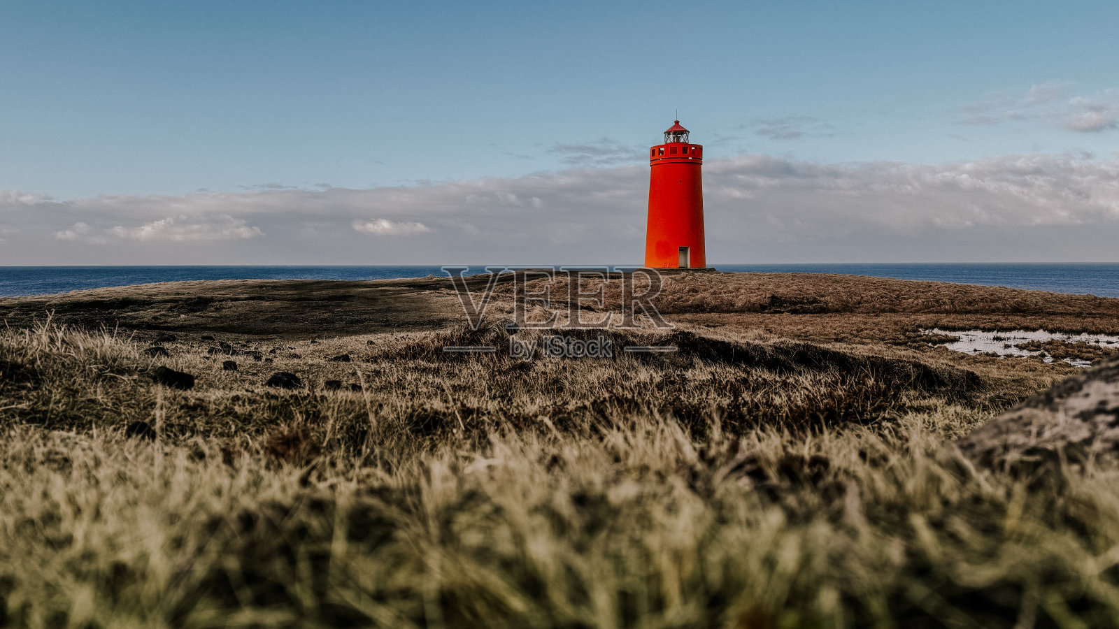 冰岛,一座红色的灯塔矗立在悬崖顶上,俯瞰着大西洋。湛蓝的天空,周围可见一些黑云,以及散射。照片摄影图片