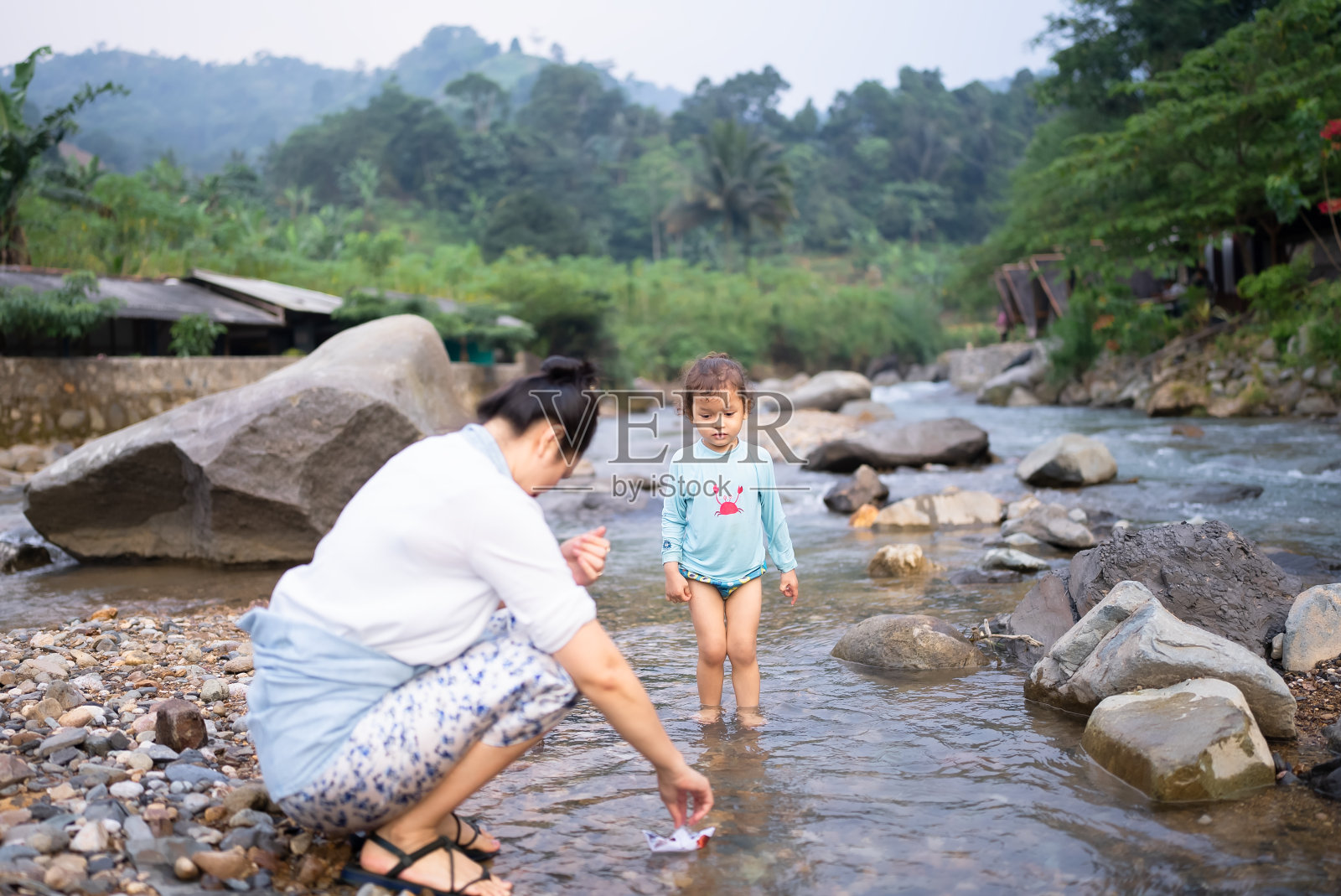 一位亚洲母亲和她的儿子把纸船放在阳光斑驳的小溪里照片摄影图片
