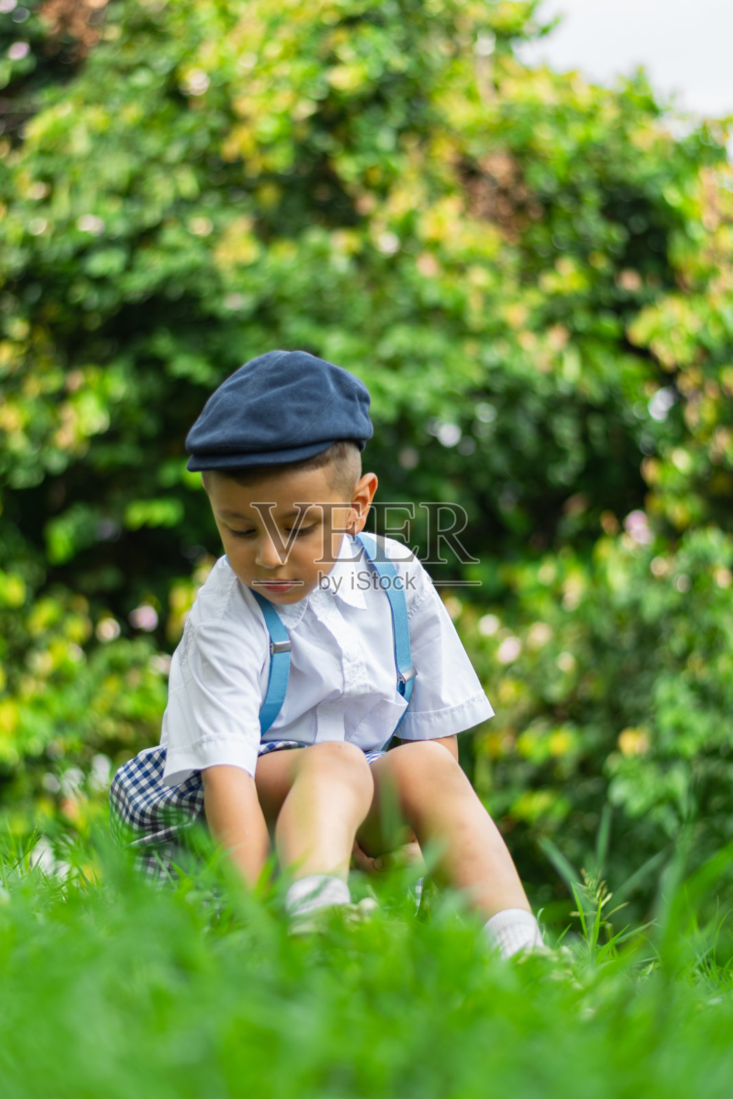 一个拉丁裔小男孩坐在公园的地上,看着他的鞋子,周围是绿色的大自然。照片摄影图片