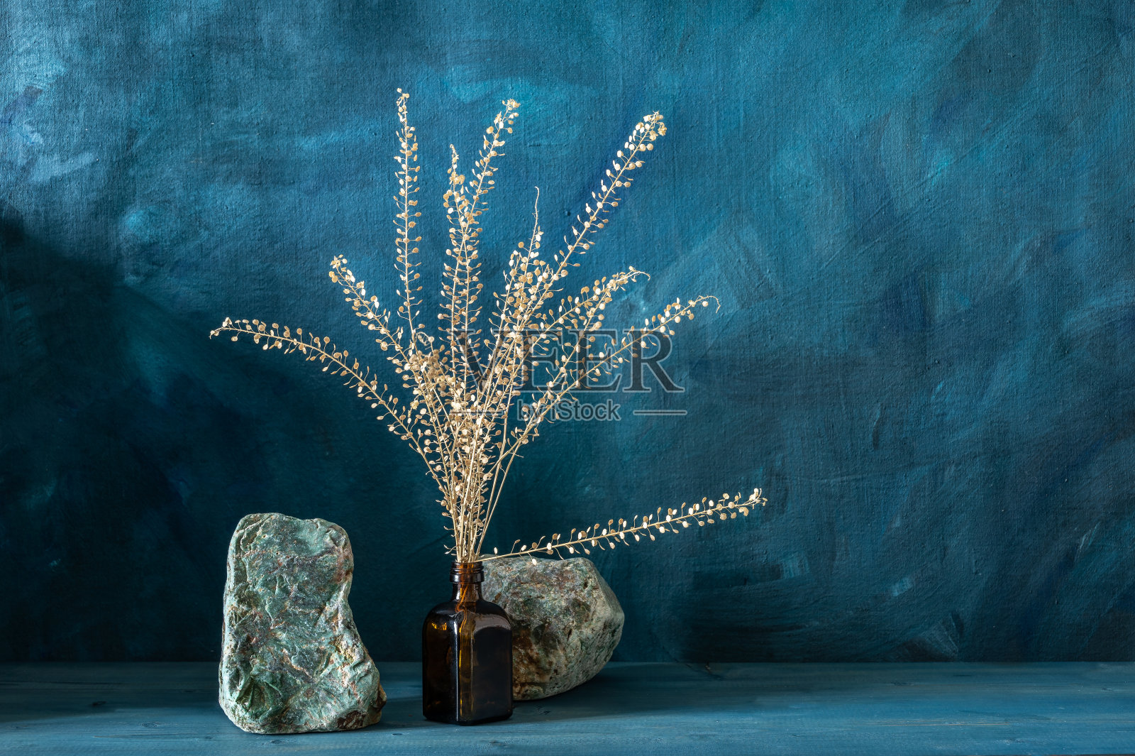 药瓶里放着干枯的花枝,蓝色的木桌上放着绿色的石头。照片摄影图片