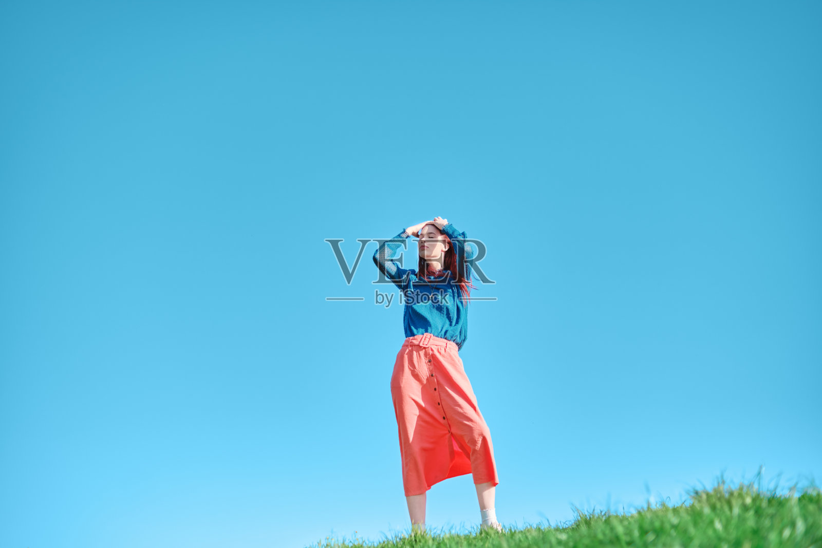 身穿蓝色上衣和珊瑚色裙子的少女站在碧绿的草地上,背景是湛蓝的天空。户外时尚摄影。夏季休闲装概念。照片摄影图片