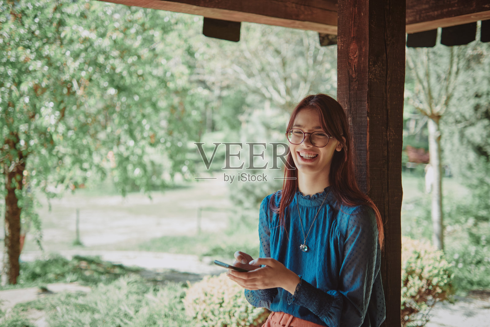 一个快乐的十几岁的女孩在一个质朴的木制露台上使用智能手机,周围是郁郁葱葱的花园。欢快的生活方式肖像,突出数字参与和户外放松。照片摄影图片