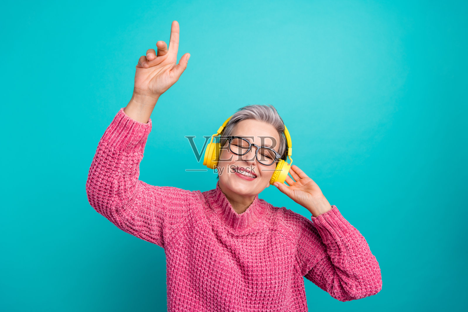 图为身穿粉色毛衣、戴着眼镜、戴着耳机欣赏音乐的女子,在蓝绿色的背景下,手指竖起照片摄影图片
