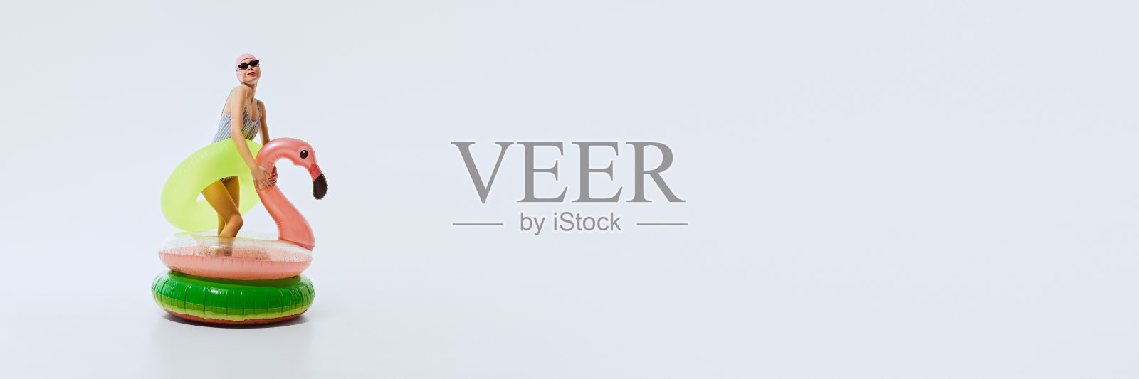 时尚的年轻女子在游泳帽和泳衣站在火烈鸟形状游泳圈内,戴着太阳镜对白色背景照片摄影图片