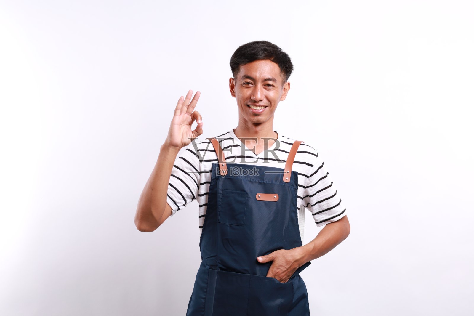 自信帅气的服务员,戴着蓝色围裙,站在白色的背景下,表示可以。照片摄影图片