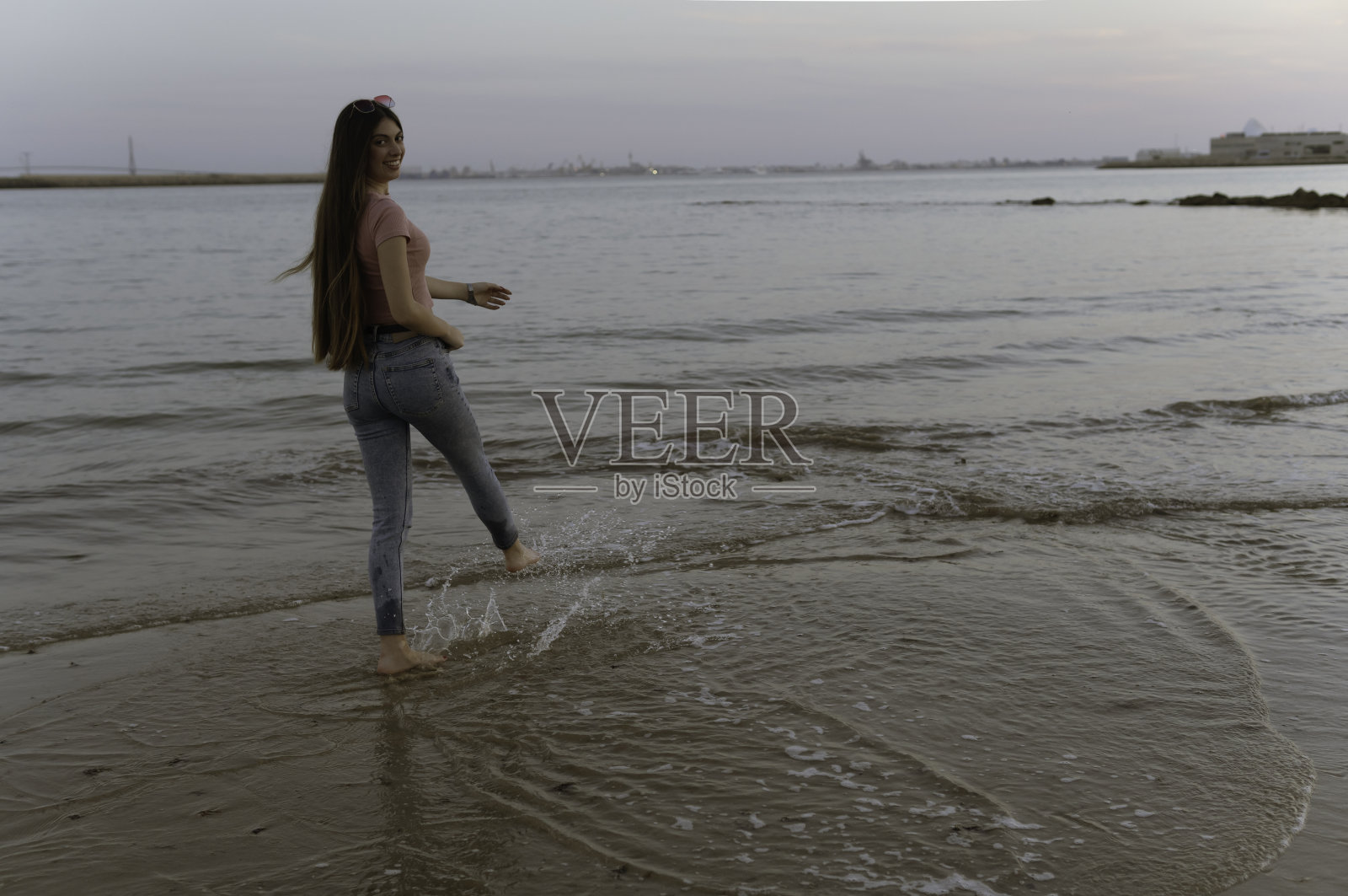 岸边的一个女人一边看着镜头,一边用脚在水里玩耍。照片摄影图片