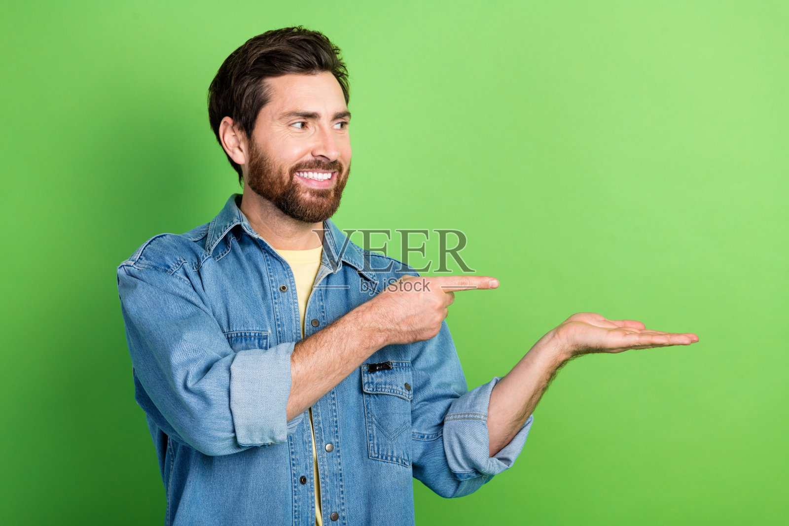 照片中心情不错的帅哥穿着牛仔夹克,手持掌心指指空白空间孤立的绿色背景照片摄影图片