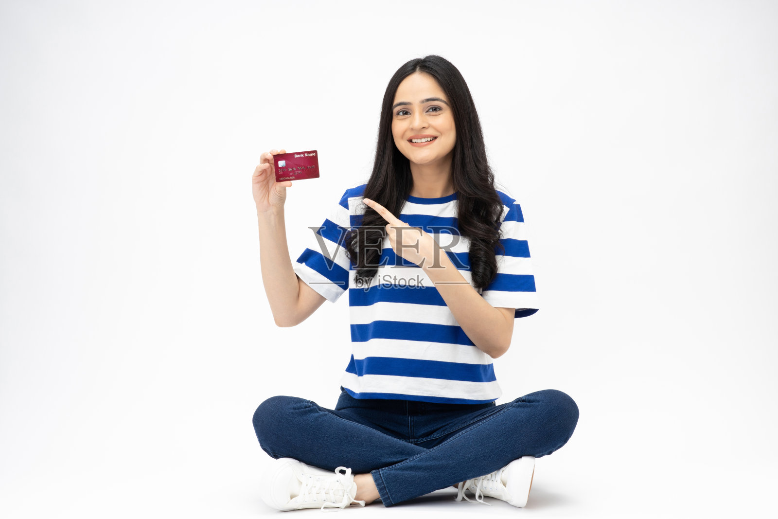 漂亮的年轻女孩的全尺寸照片,坐在地板上,显示信用卡,穿着时髦的衣服,孤立在白色背景的股票照片照片摄影图片