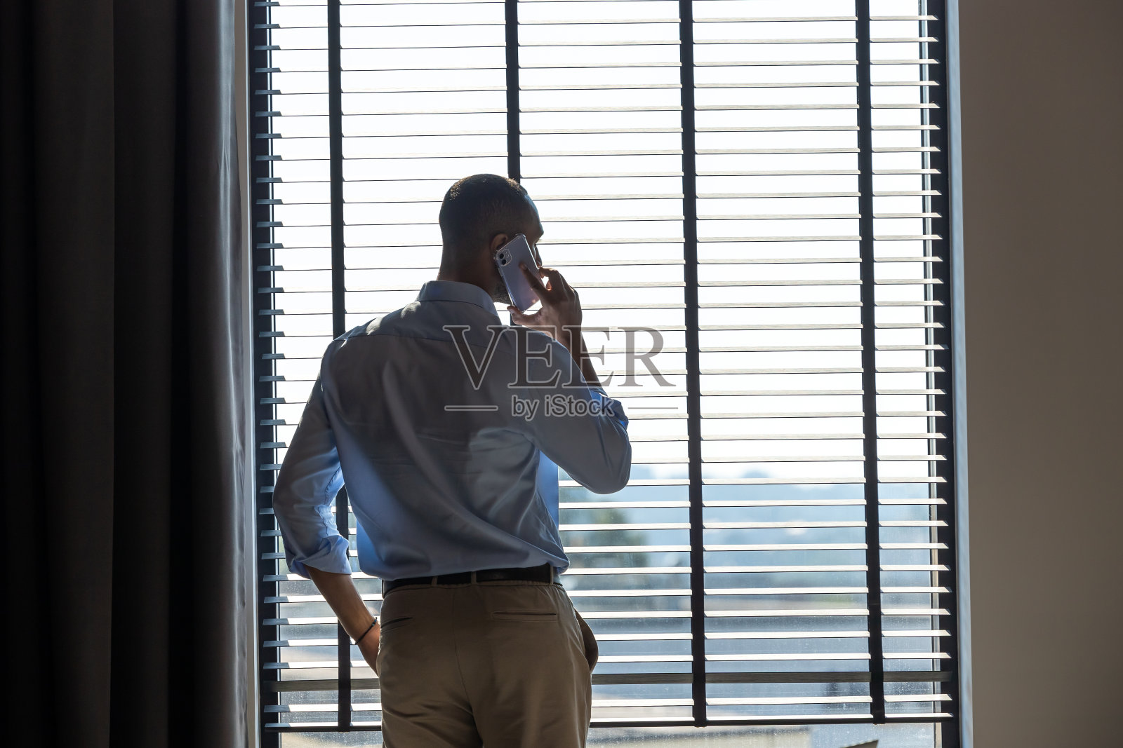 商人站在办公室的大窗户旁边,自信地讲着手机照片摄影图片