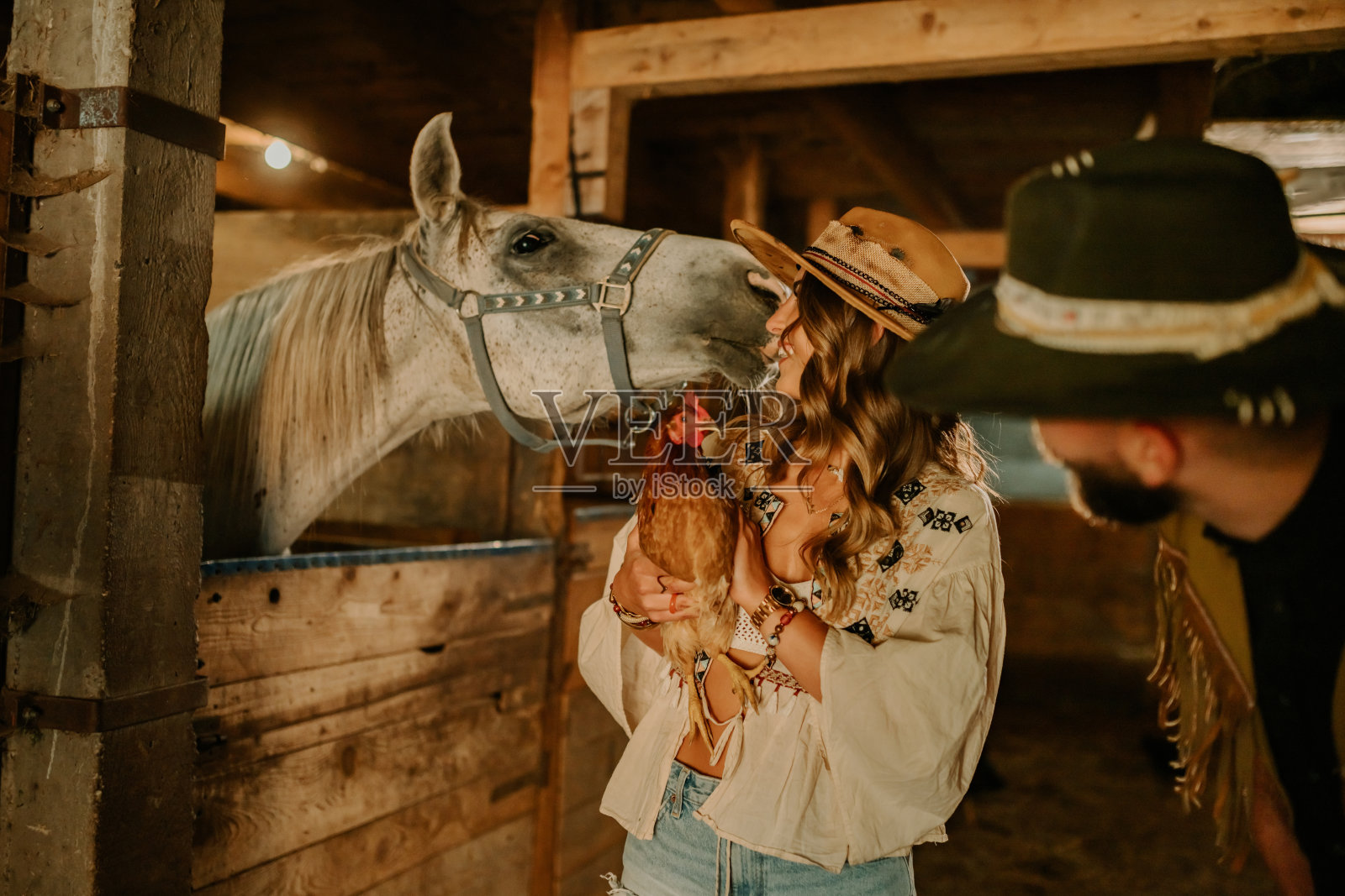 兴高采烈的女牛仔抱着一只母鸡,对着马厩里的马微笑。照片摄影图片