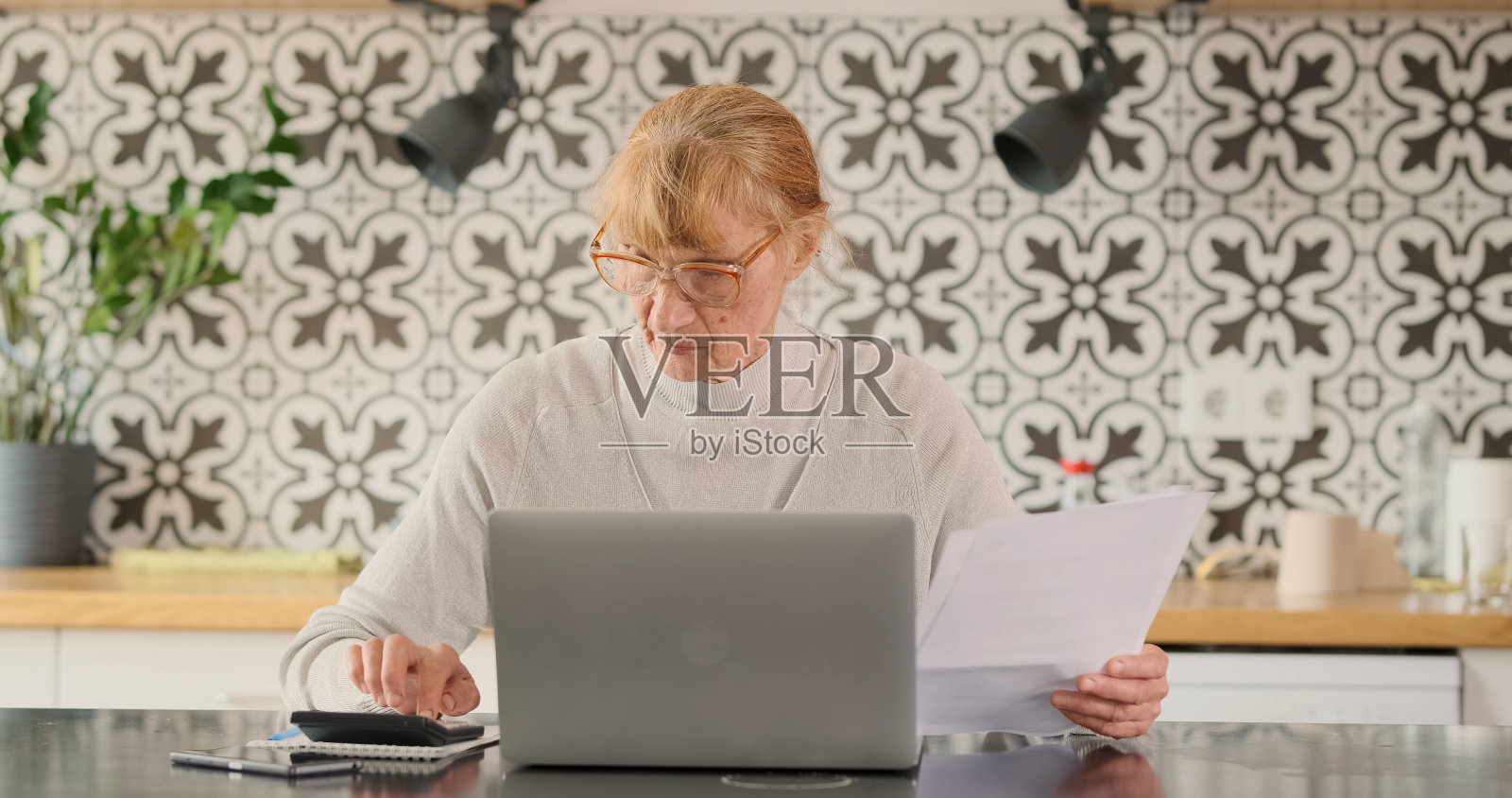 老妇人拿着纸质的服务账单,在计算器、笔记本电脑、桌面厨房上计算费用。肖像,正面。纳税、计算费用、省钱的观念照片摄影图片