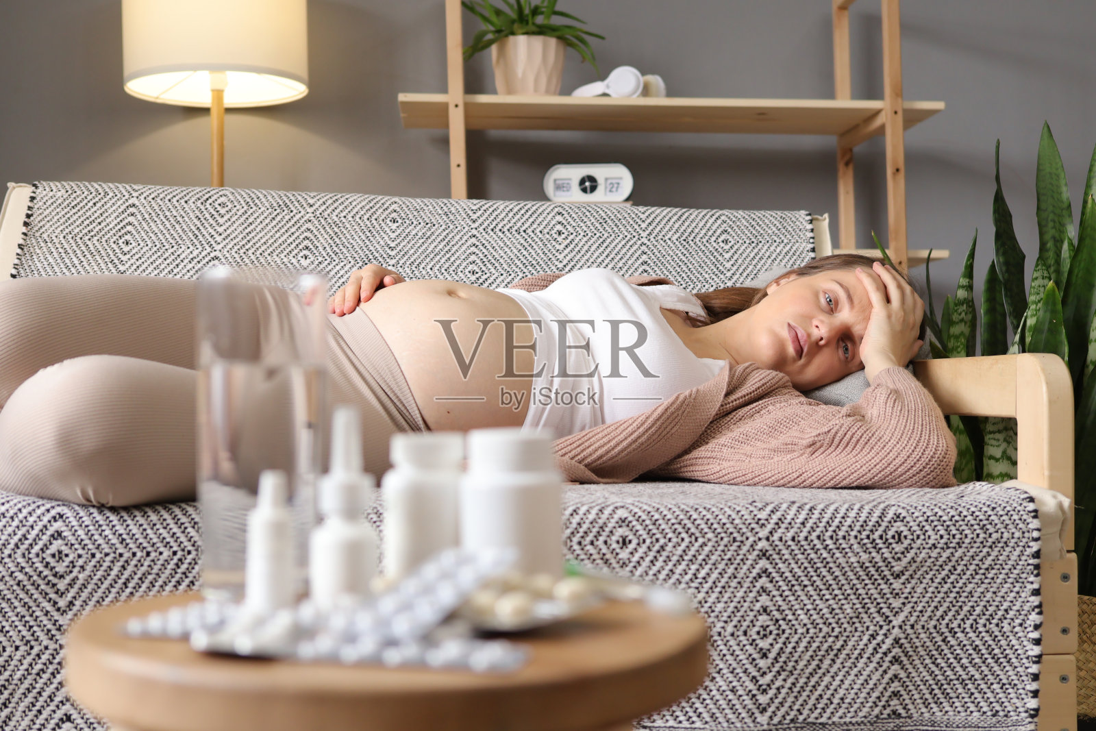 生病的孕妇躺在家里的沙发上,背景是各种各样的药丸,不健康的女性患流感和高温触摸疼痛的头部照片摄影图片