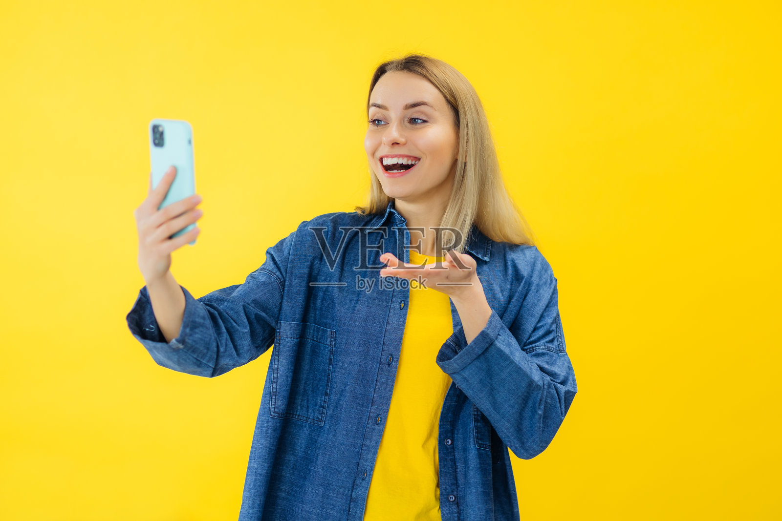 漂亮的年轻女士穿着休闲牛仔衬衫,在黄色的工作室背景上用现代手机进行视频通话。照片摄影图片