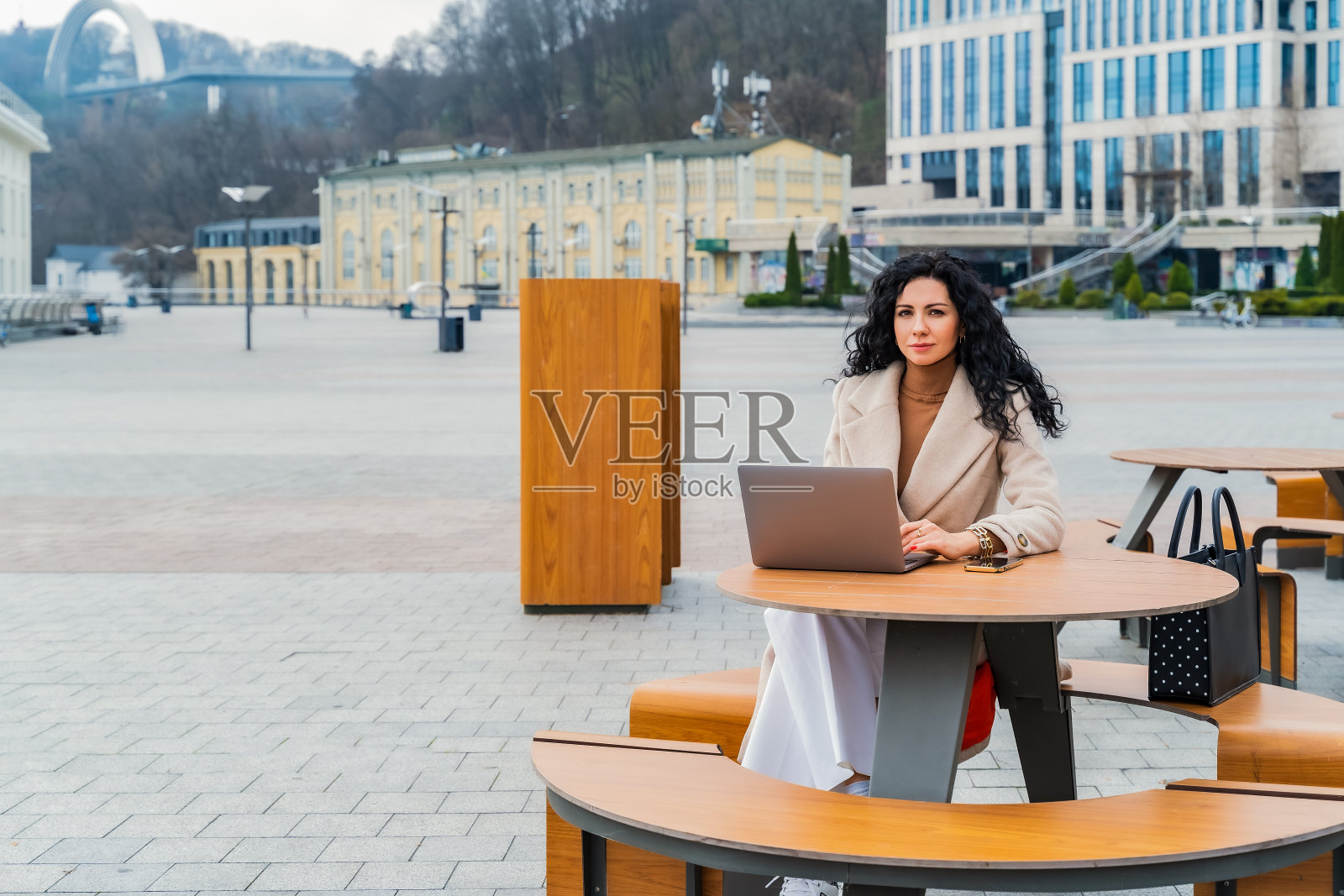 女老板坐在城市街道上的一张木桌旁,外套里装着一台笔记本电脑照片摄影图片