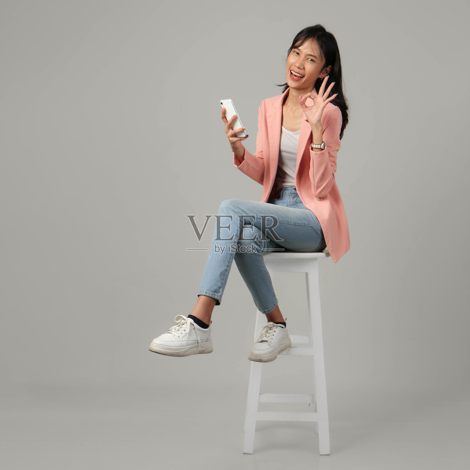 在孤立的背景下,快乐兴奋的亚洲女性坐在凳子上,拿着手机,握紧拳头照片摄影图片