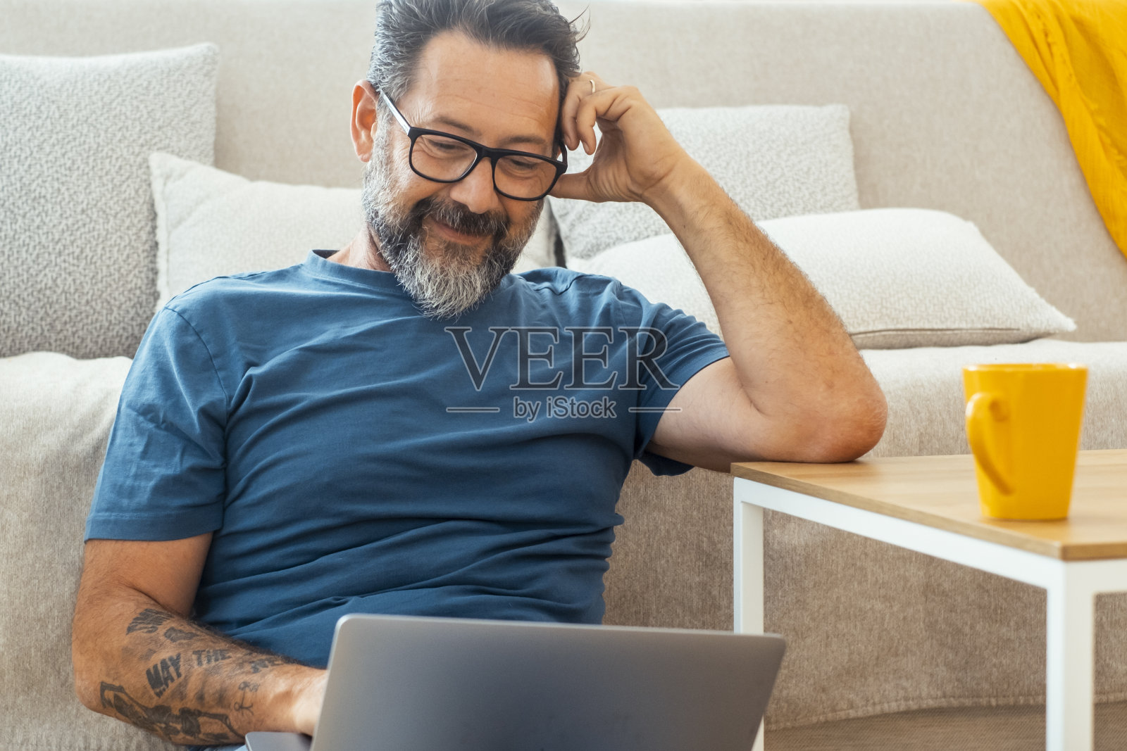 穿着休闲服,戴着眼镜的成年男子,用着笔记本电脑,微笑着坐在家里的沙发上。室内科技电脑休闲活动人们的生活方式。在线工作照片摄影图片