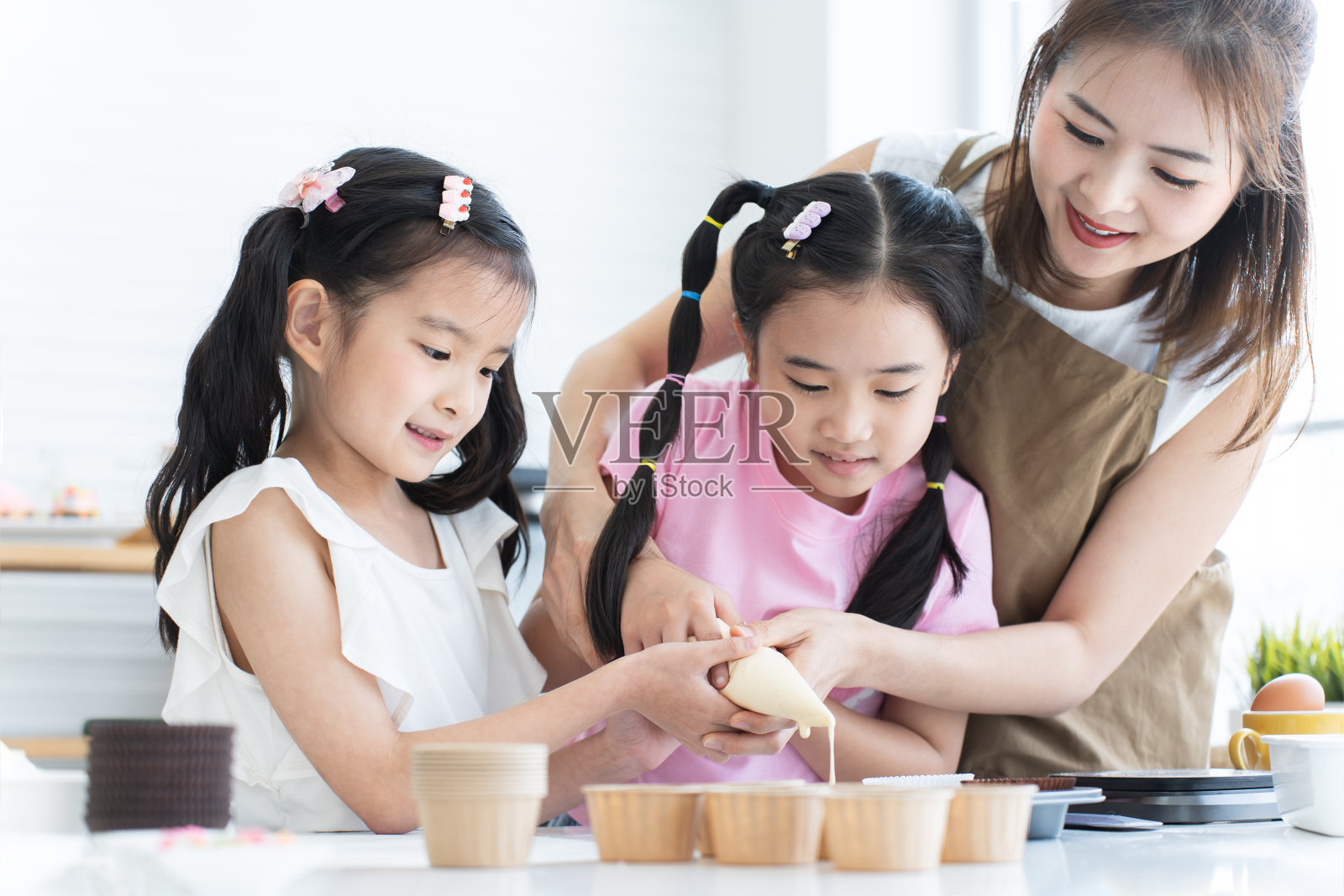 一个亚洲家庭,母亲和两个女儿喜欢在厨房里一起做纸杯蛋糕。妈妈和小女孩把蛋糕糊挤进纸杯蛋糕纸杯里,准备烘烤,微笑着。节日快乐照片摄影图片