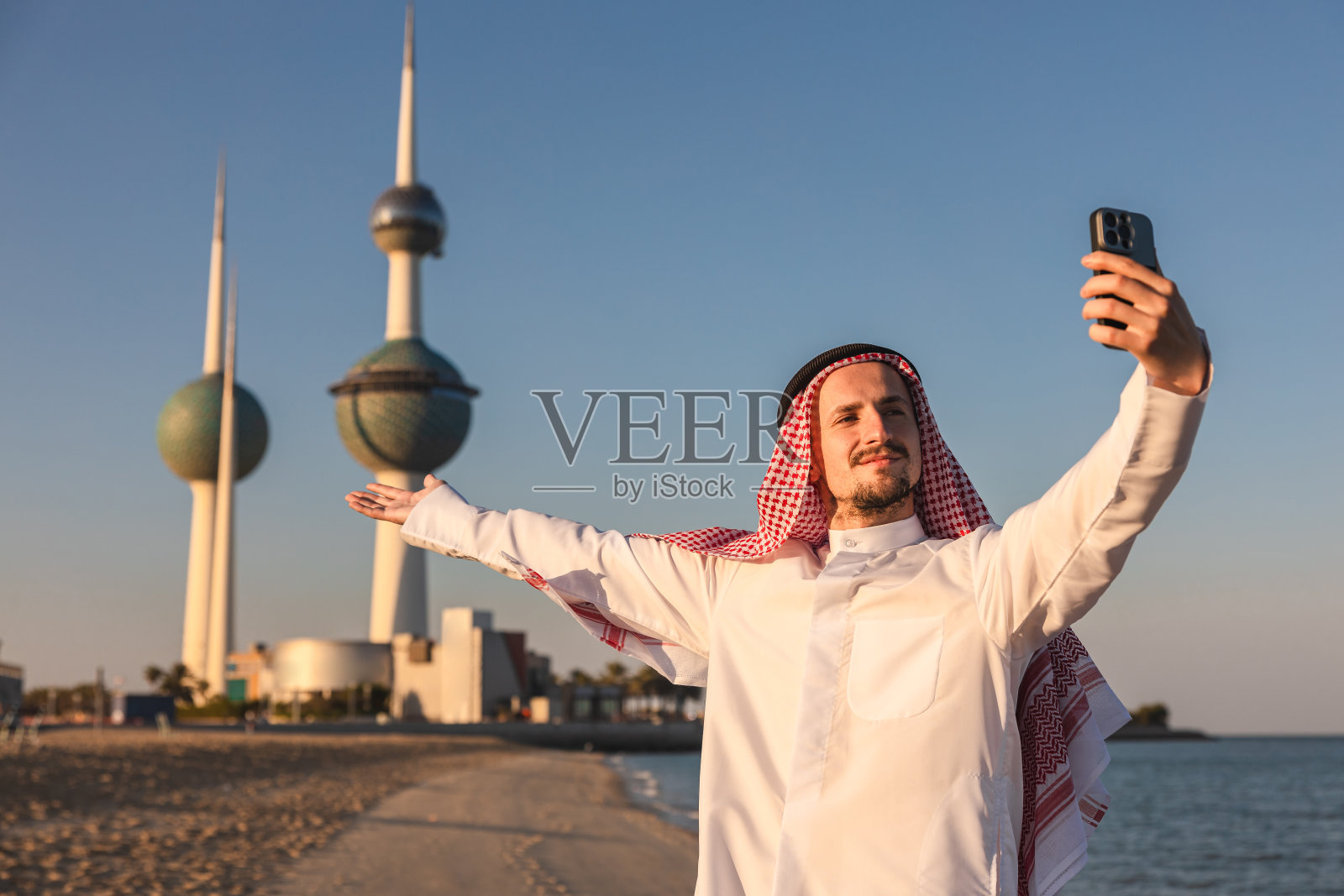 在海边使用手机的年轻酋长,背景是科威特塔照片摄影图片