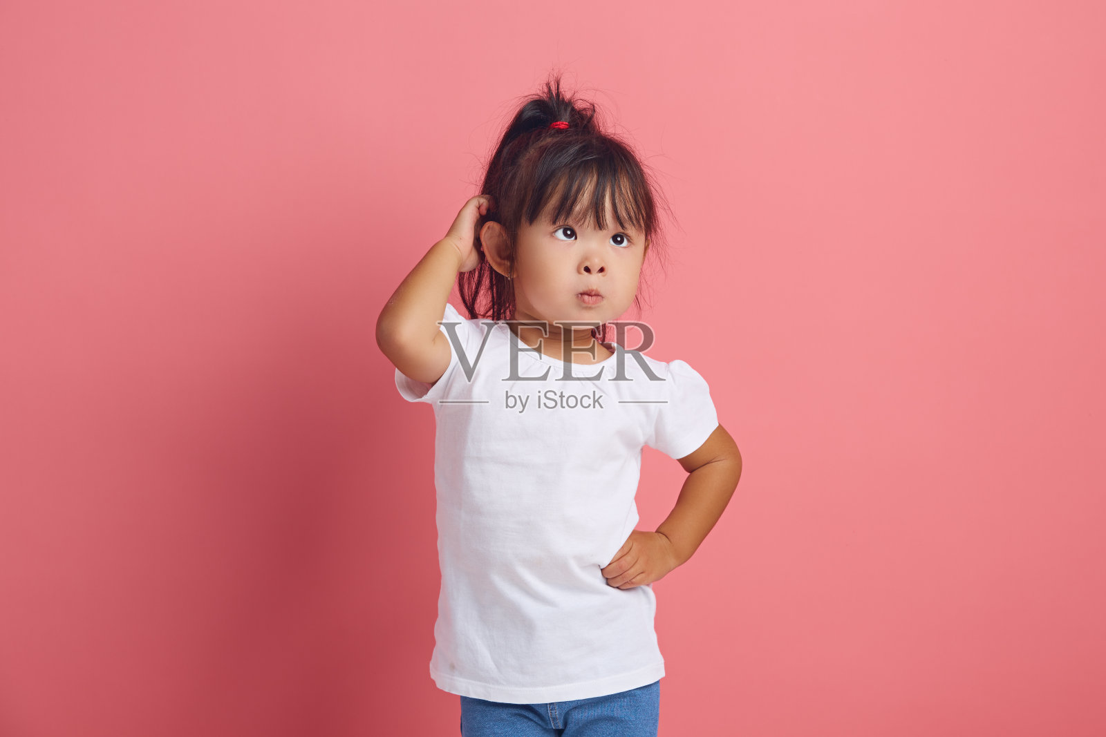 困惑的小女孩用沉思的眼神挠着头,脸上带着困惑的表情,穿着中性的白色t恤,在粉红色的背景下摆出困惑的姿势。照片摄影图片