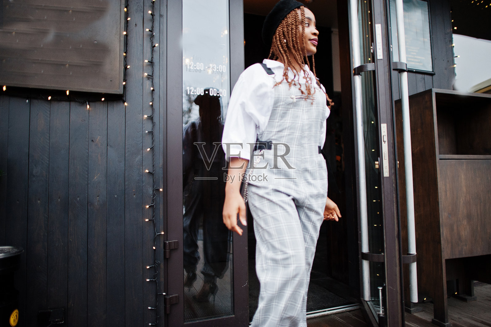 穿着工装裤和贝雷帽的非裔美国妇女在装饰着圣诞装饰的户外露台上走出门。照片摄影图片