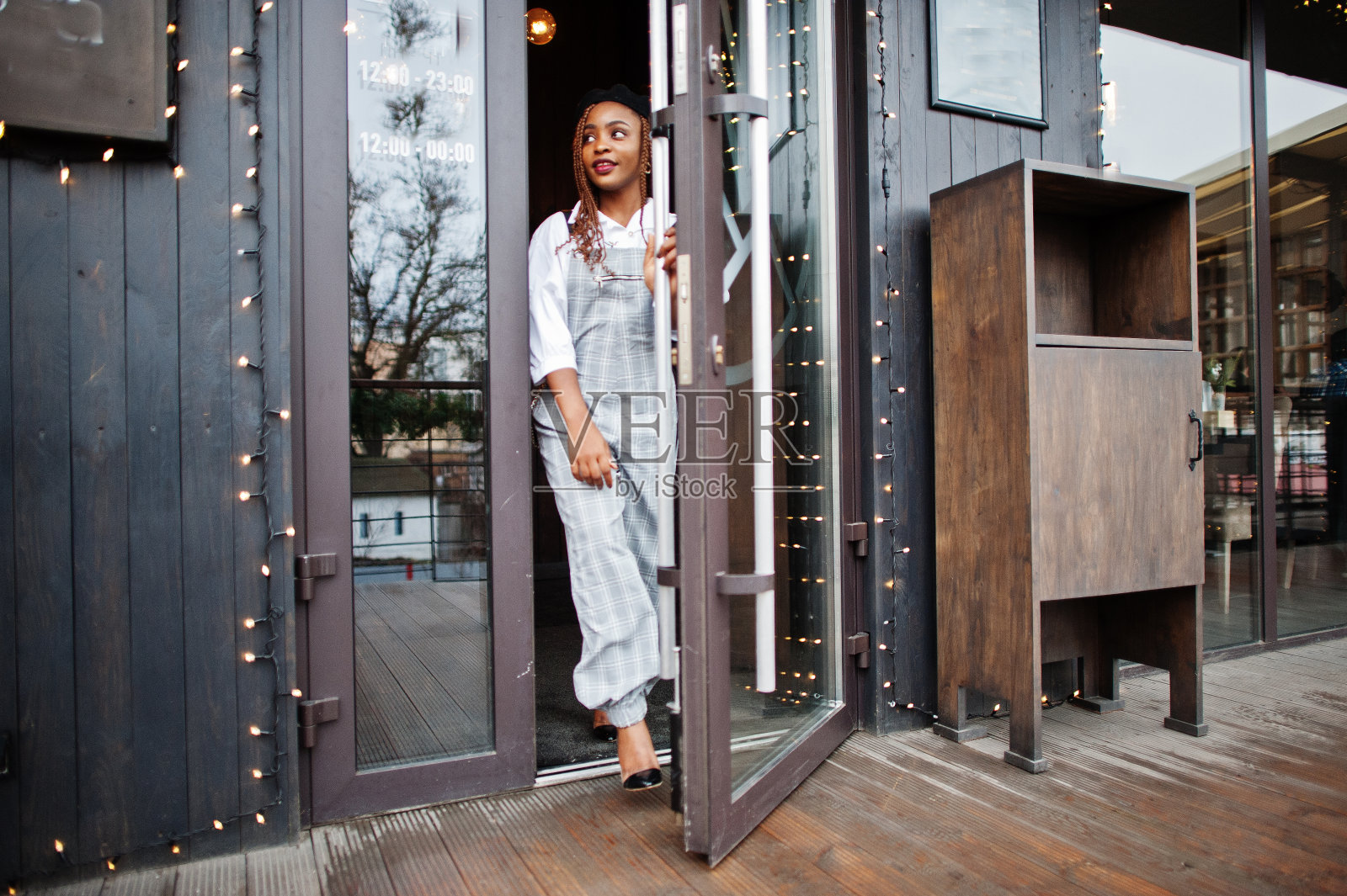 穿着工装裤和贝雷帽的非裔美国妇女在装饰着圣诞装饰的户外露台上走出门。照片摄影图片