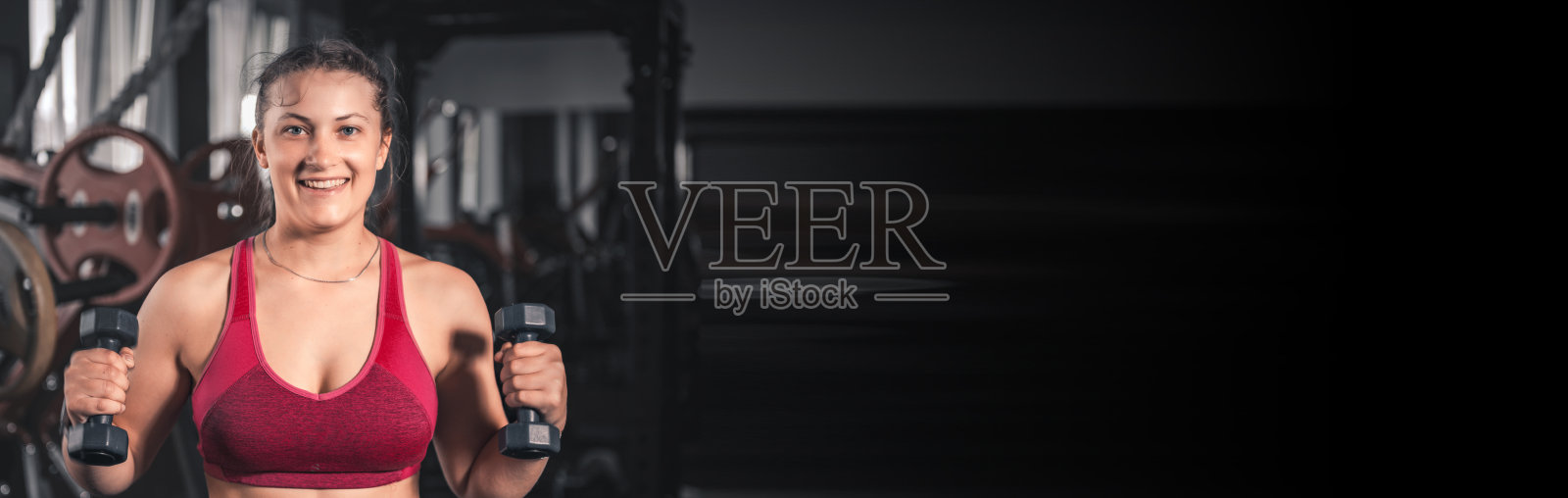年轻女子在健身房拿着哑铃。穿着运动服的年轻迷人女子手持哑铃进行健身锻炼的肖像照片摄影图片