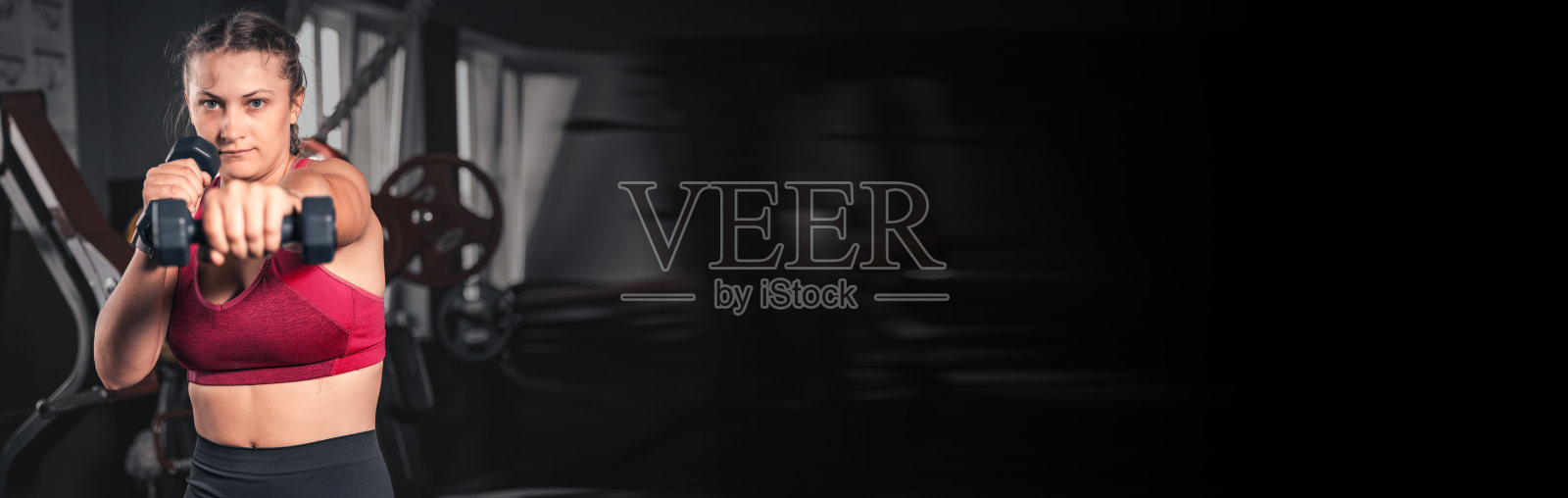 年轻女子在健身房拿着哑铃。穿着运动服的年轻迷人女子手持哑铃进行健身锻炼的肖像照片摄影图片
