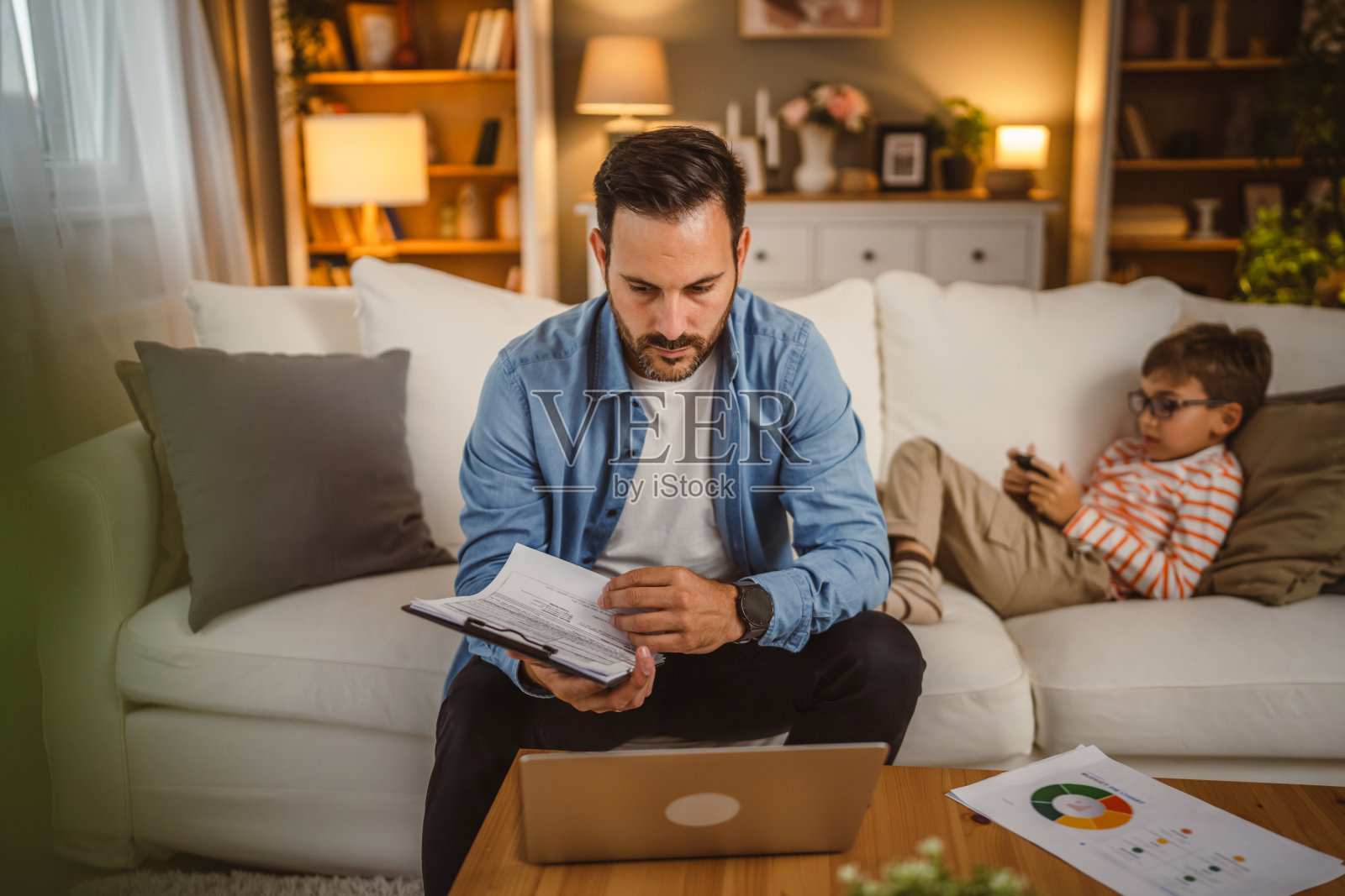 父亲在家工作,手里拿着文件和笔记本电脑,儿子在后台照片摄影图片