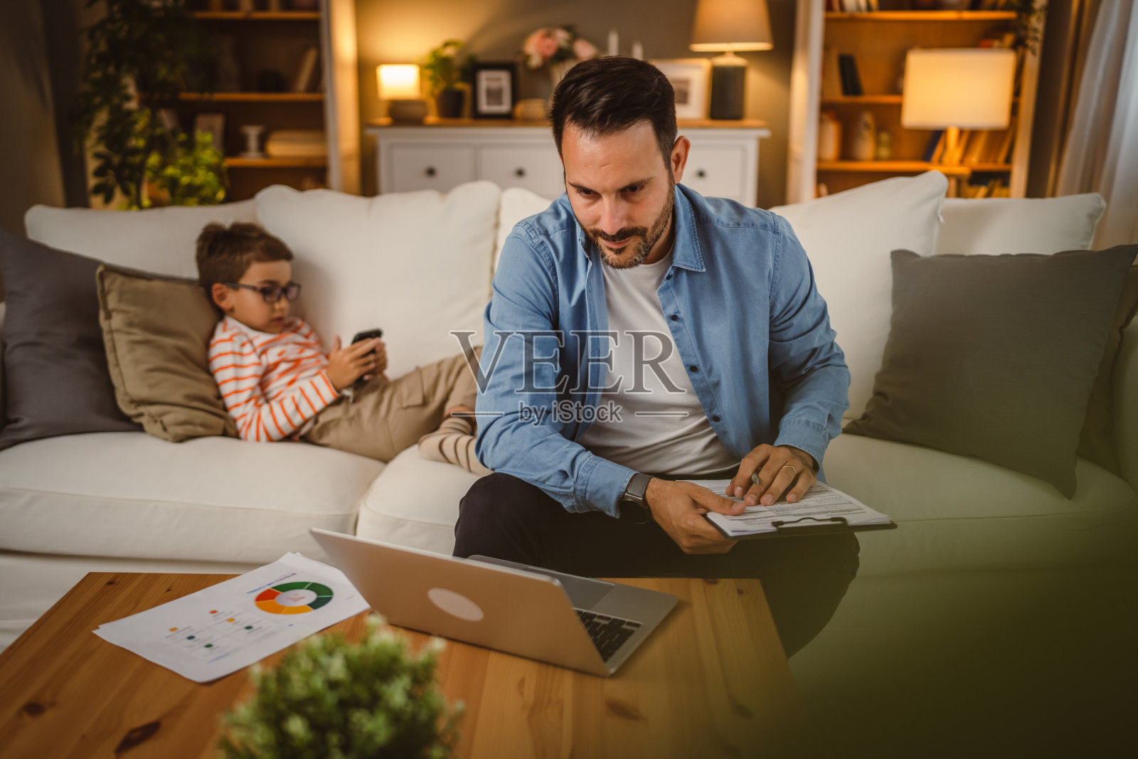 父亲在家工作,手里拿着文件和笔记本电脑,儿子在后台照片摄影图片