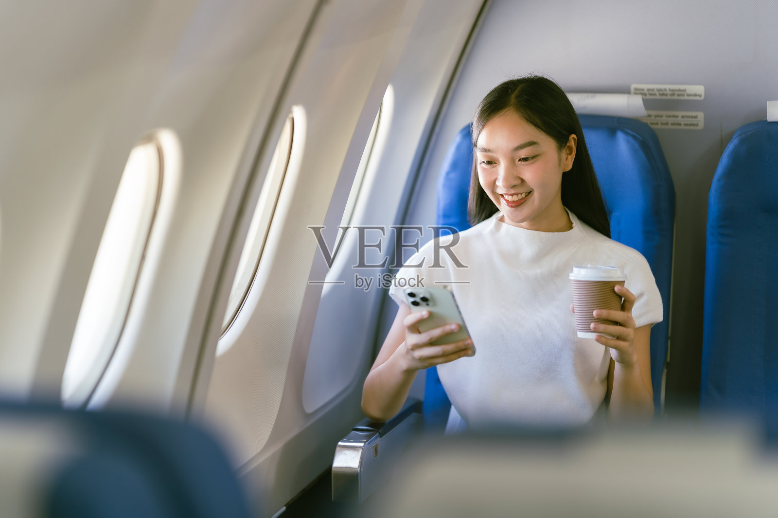 年轻的亚洲女性坐在飞机上用智能手机聊天,坐在经济舱靠窗的座位上,在飞机上使用智能手机照片摄影图片