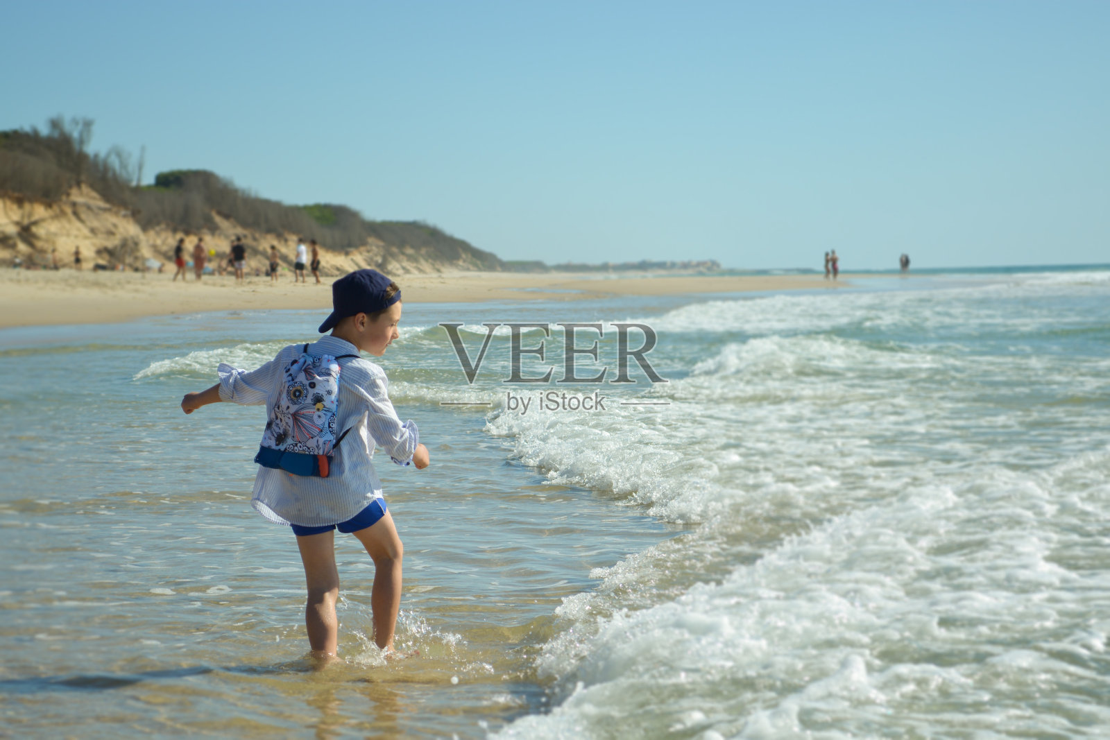 在海边,一个头戴帽子、身穿衬衫的蹒跚学步的男孩用手指着海里的什么东西。照片摄影图片