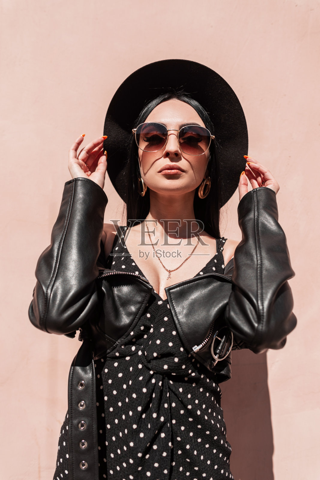 漂亮时尚的年轻女子,戴着帽子,穿着夹克,穿着裙子,戴着太阳镜,在城市里享受阳光。阳光肖像时尚女孩模特在黑色优雅的夏天美丽的衣服在明亮的阳光下靠近粉红色的墙照片摄影图片