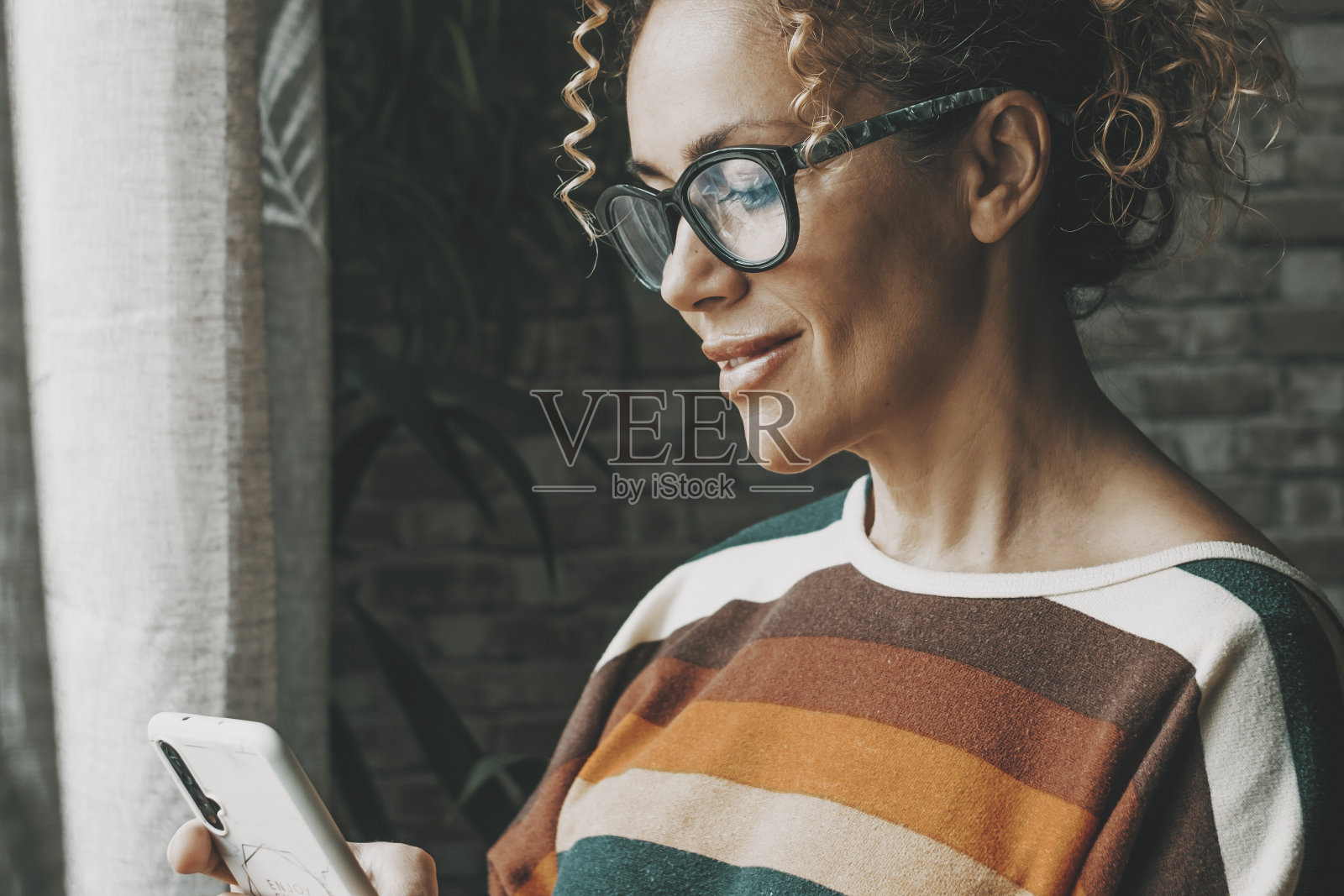 一个快乐的女人在室内对着手机微笑,用应用程序聊天或给人发信息。约会和孤独的生活方式与科技和互联网连接的女性。年轻女士在手机上写字照片摄影图片