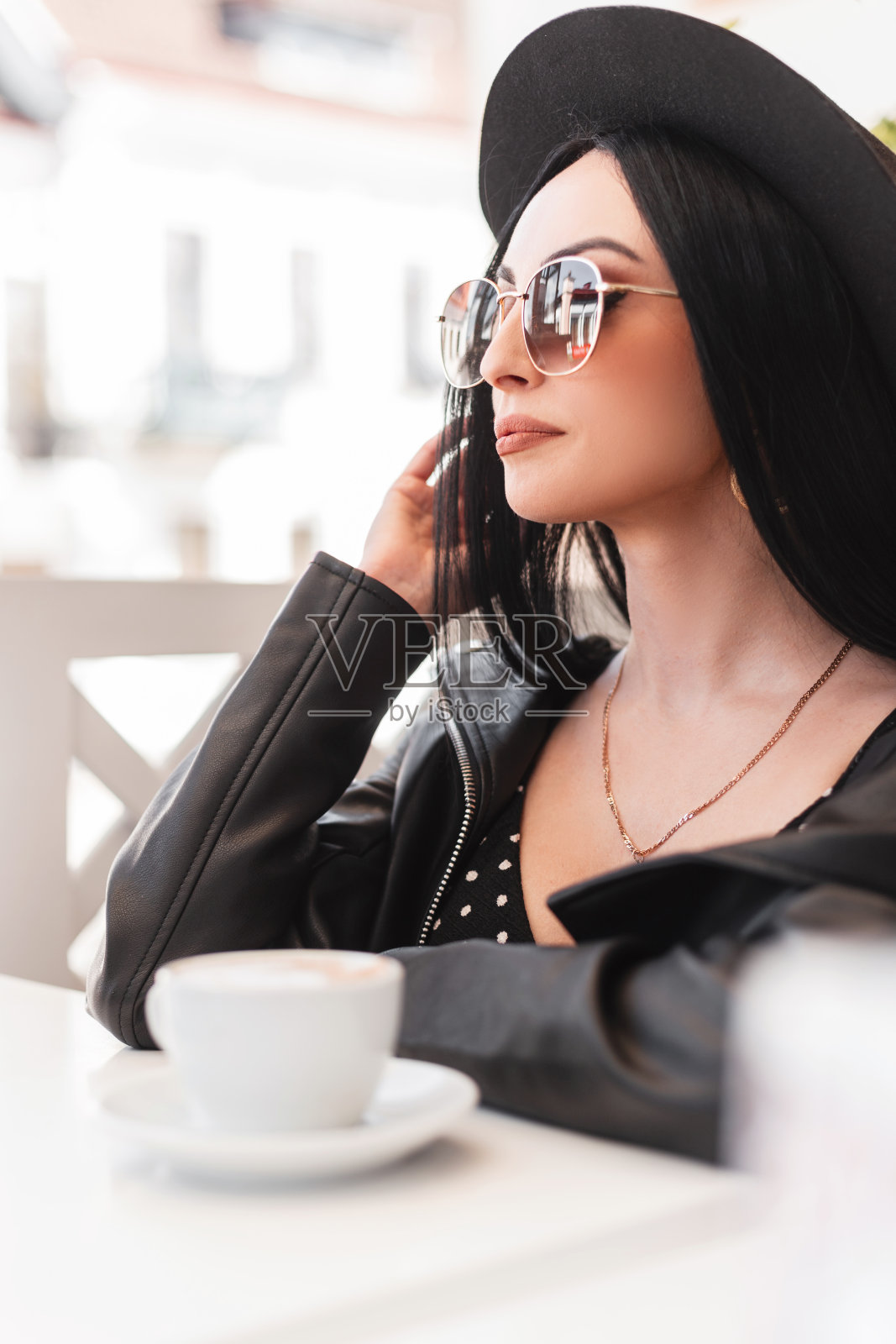 一个可爱的时尚美女,戴着太阳镜和帽子,穿着时尚的皮夹克和裙子,坐在咖啡馆里喝着美味的咖啡照片摄影图片